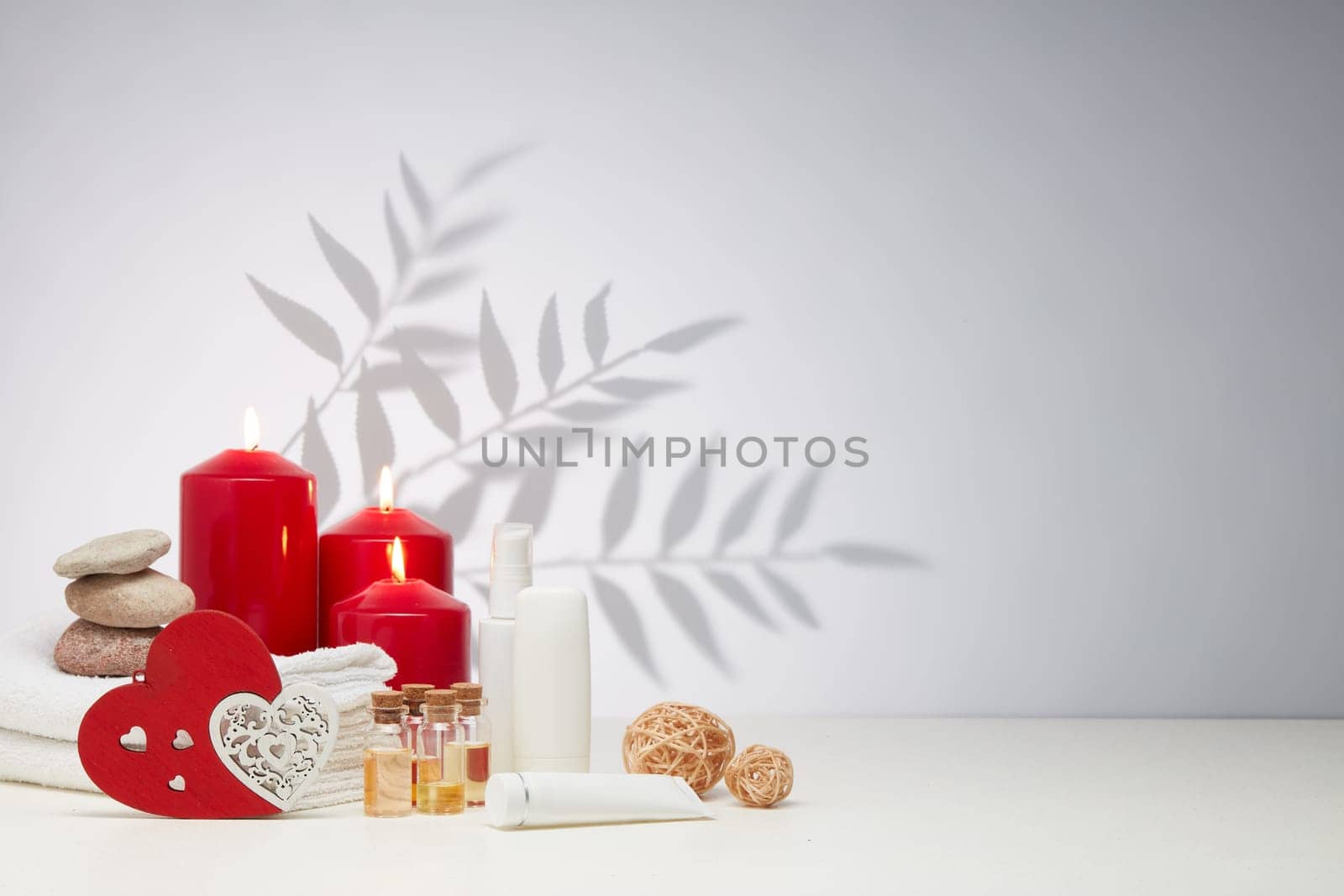 Spa still life with creams, essential oils, burning red candles, hearts on light background with shadow from plant. Healthy lifestyle, body care, Spa treatment. Valentine's Day. Honeymoon. Wedding