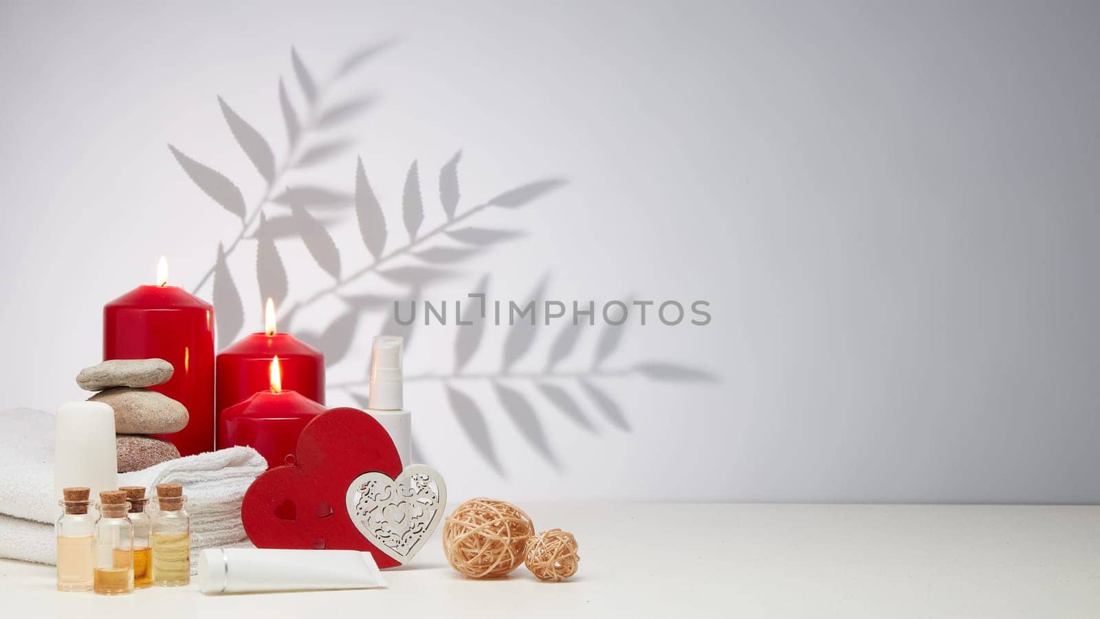 Spa still life with creams, essential oils, burning red candles, hearts on light background with shadow from plant. Healthy lifestyle, body care, Spa treatment. Valentine's Day. Honeymoon. Wedding