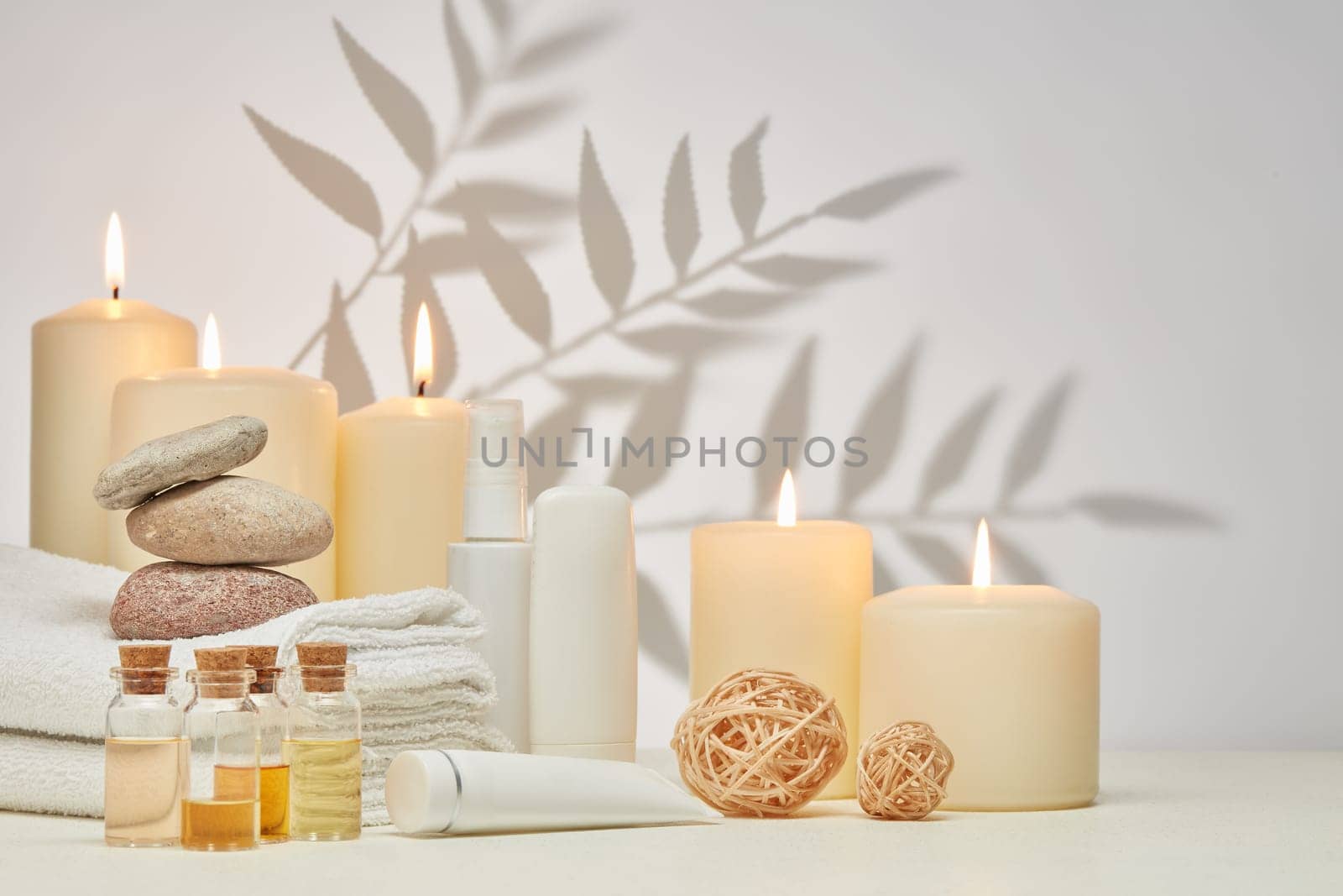 Spa still life with creams, essential oils, burning candles, stones and towels on light background with shadow from plant. Healthy lifestyle, body care, Spa treatment.