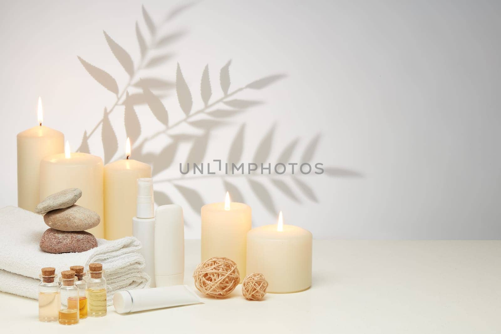 Spa still life with creams, essential oils, burning candles, stones and towels on light background with shadow from plant. Healthy lifestyle, body care, Spa treatment.
