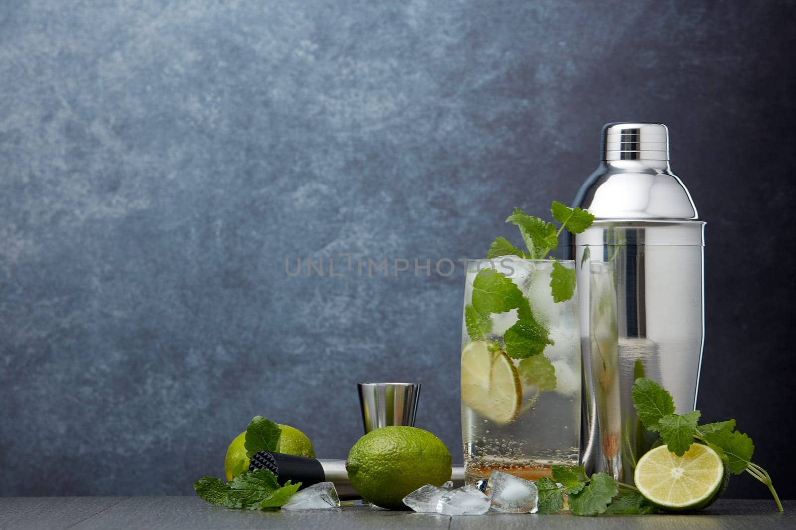Set of bar tools: cocktail shaker, muddler, jigger, cocktail with ice in highball glass,  lime and mint on dark gray background with copy space. Bartender's tool. Mojito cocktail making.