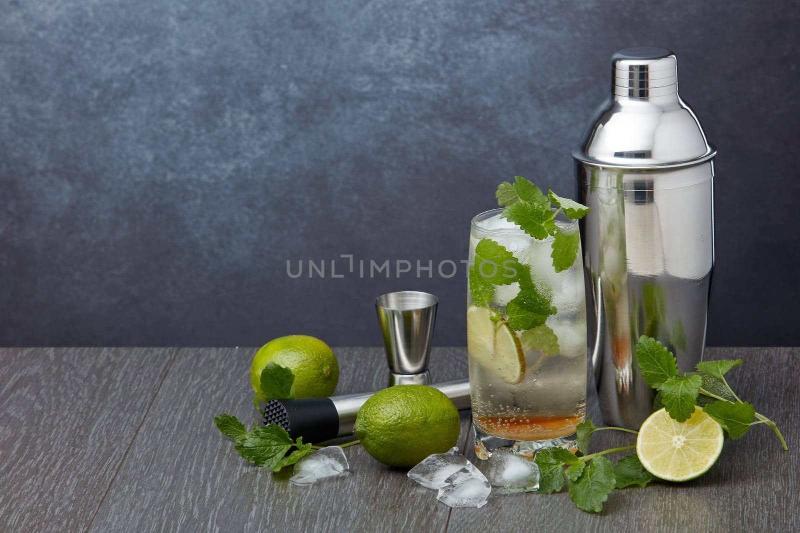 Set of bar tools: cocktail shaker, muddler, jigger, cocktail with ice in highball glass,  lime and mint on dark gray background with copy space. Bartender's tool. Mojito cocktail making.