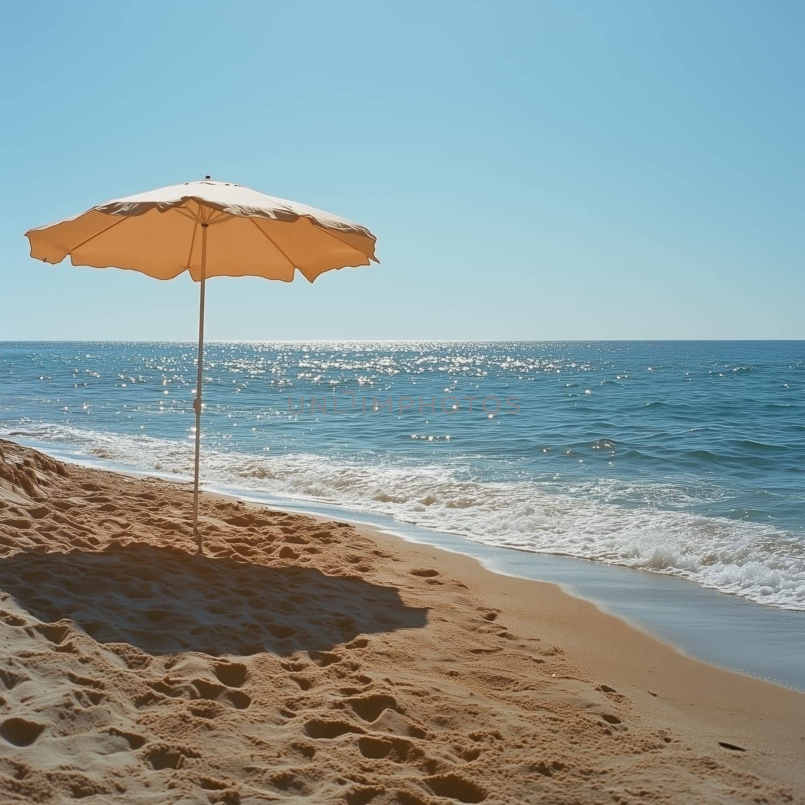 A single beige beach umbrella stands on golden sand near the shoreline, with gentle ocean waves and a bright blue sky in the background.