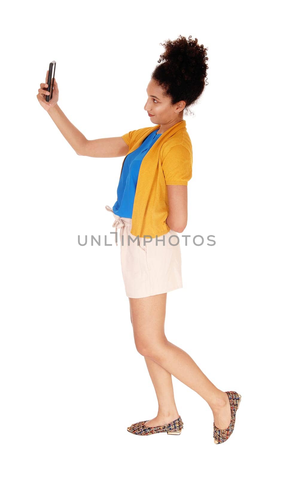 Beautiful young multi-racial woman standing in shorts and a yellow.sweater in profile looking at cellphone isolated for white background.