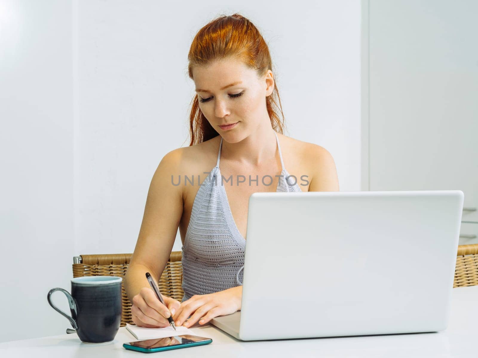 Beautiful young woman with red hair sitting with a laptop and writing on a notepad.