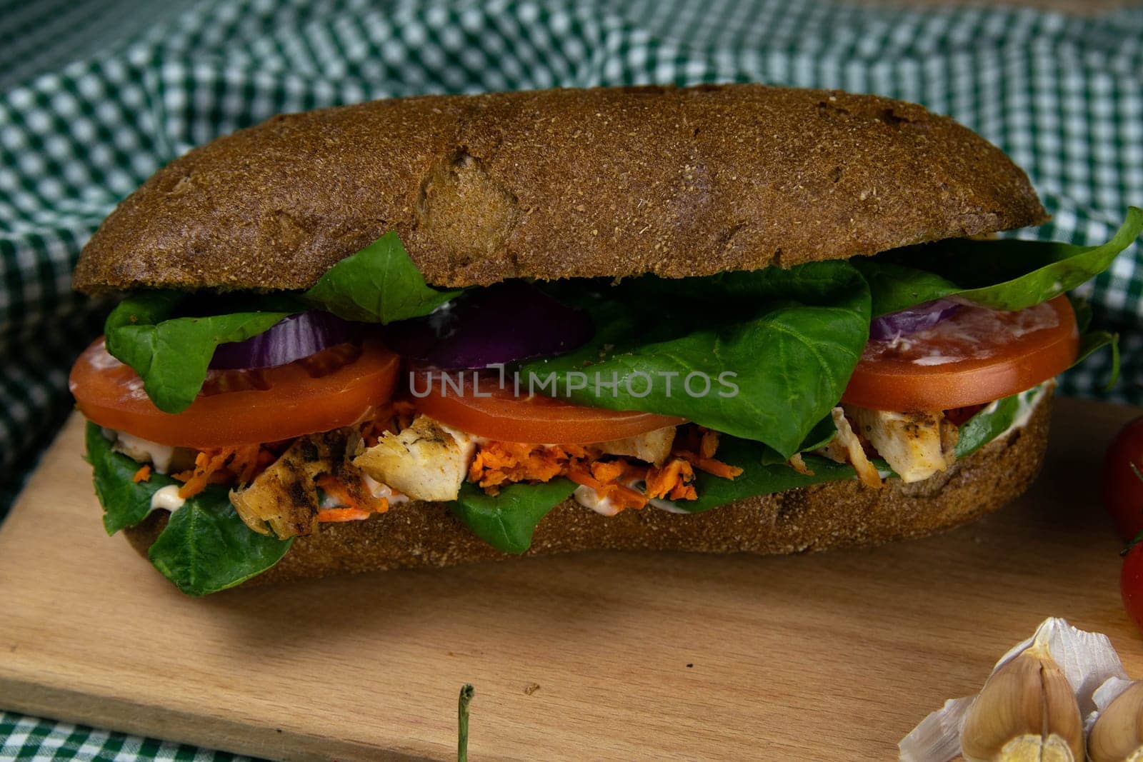 A dark bread sandwich ut in half with chicken, tomatoes, spinach, cheese and carrots stands on a wooden board that stands on a checkered white green towel. Top views