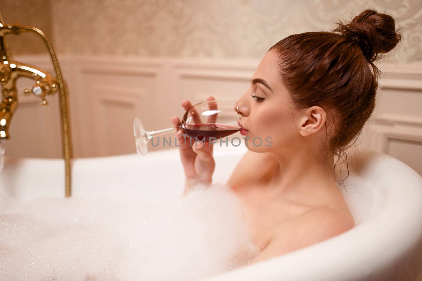 Beautiful nude woman drinking red wine in the bathtub with foam.