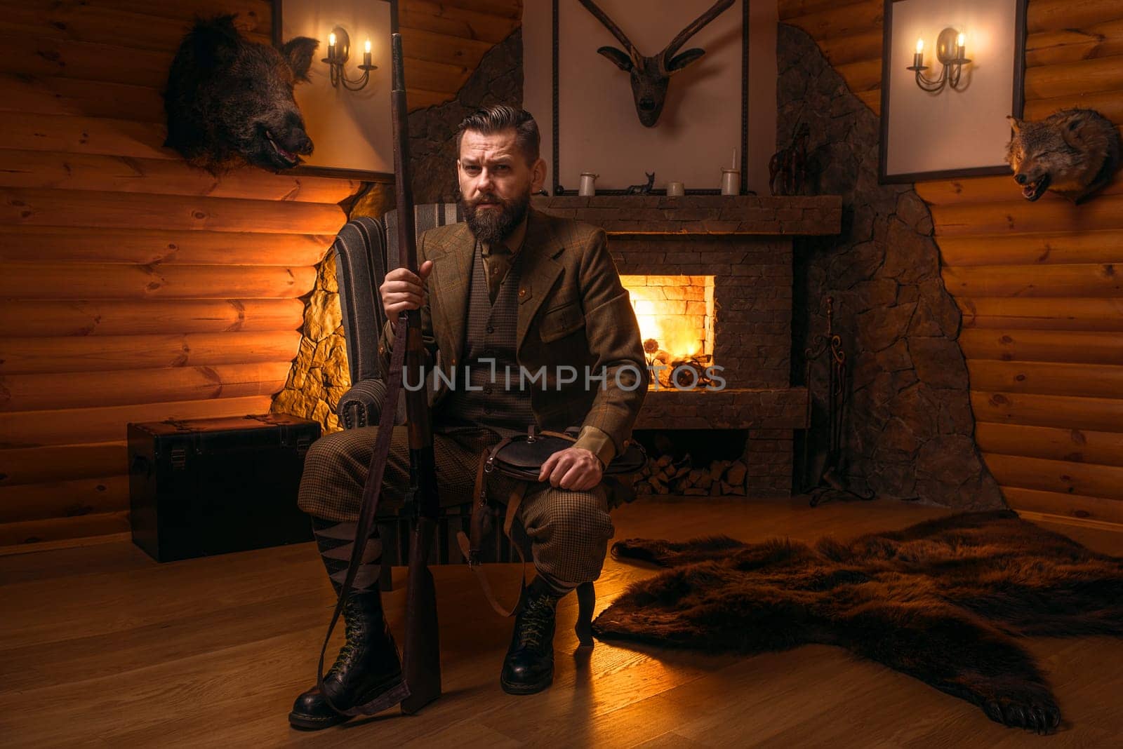 Vintage hunter man in traditional hunting clothing sitting in a chair with retro rifle against burning fireplace. Stuffed wild animals, bear skin and other trophies on background