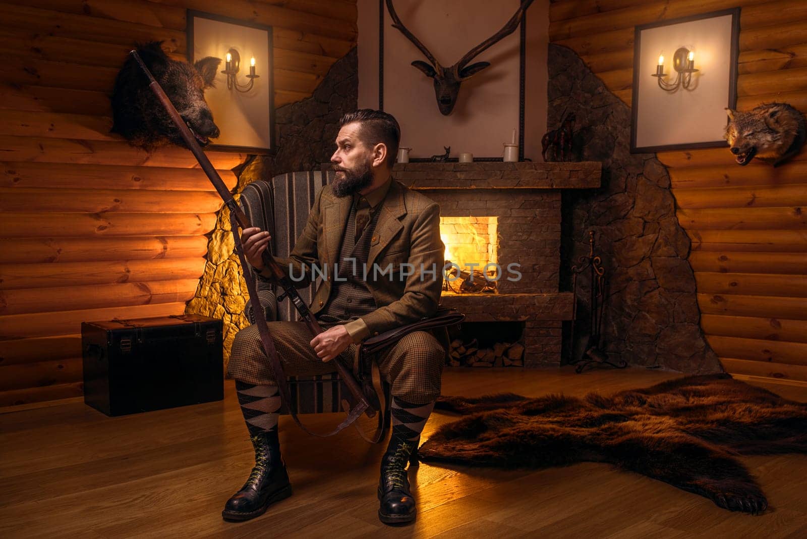 Vintage hunter man in traditional hunting clothing sitting in a chair with retro rifle against burning fireplace. Stuffed wild animals, bear skin and other trophies on background
