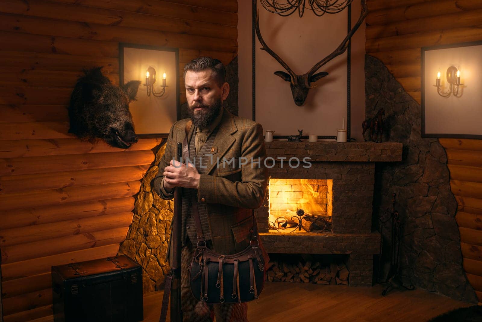 Hunter man with old gun in vintage traditional hunting clothing standing against antique chest. Fireplace, stuffed wild animals, bear skin and other trophies on background