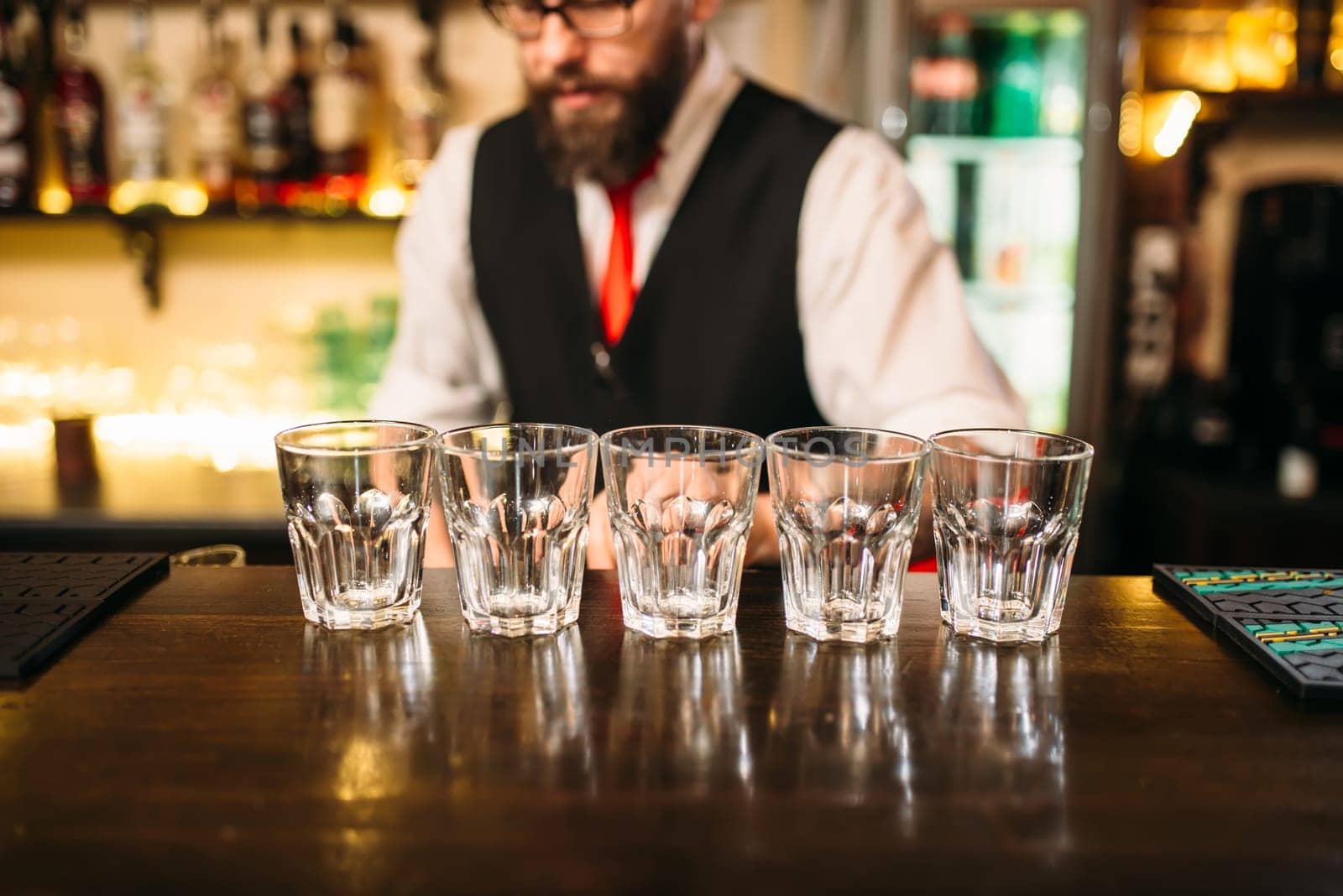 Empty glasses on wooden bar counter, bartender working on background
