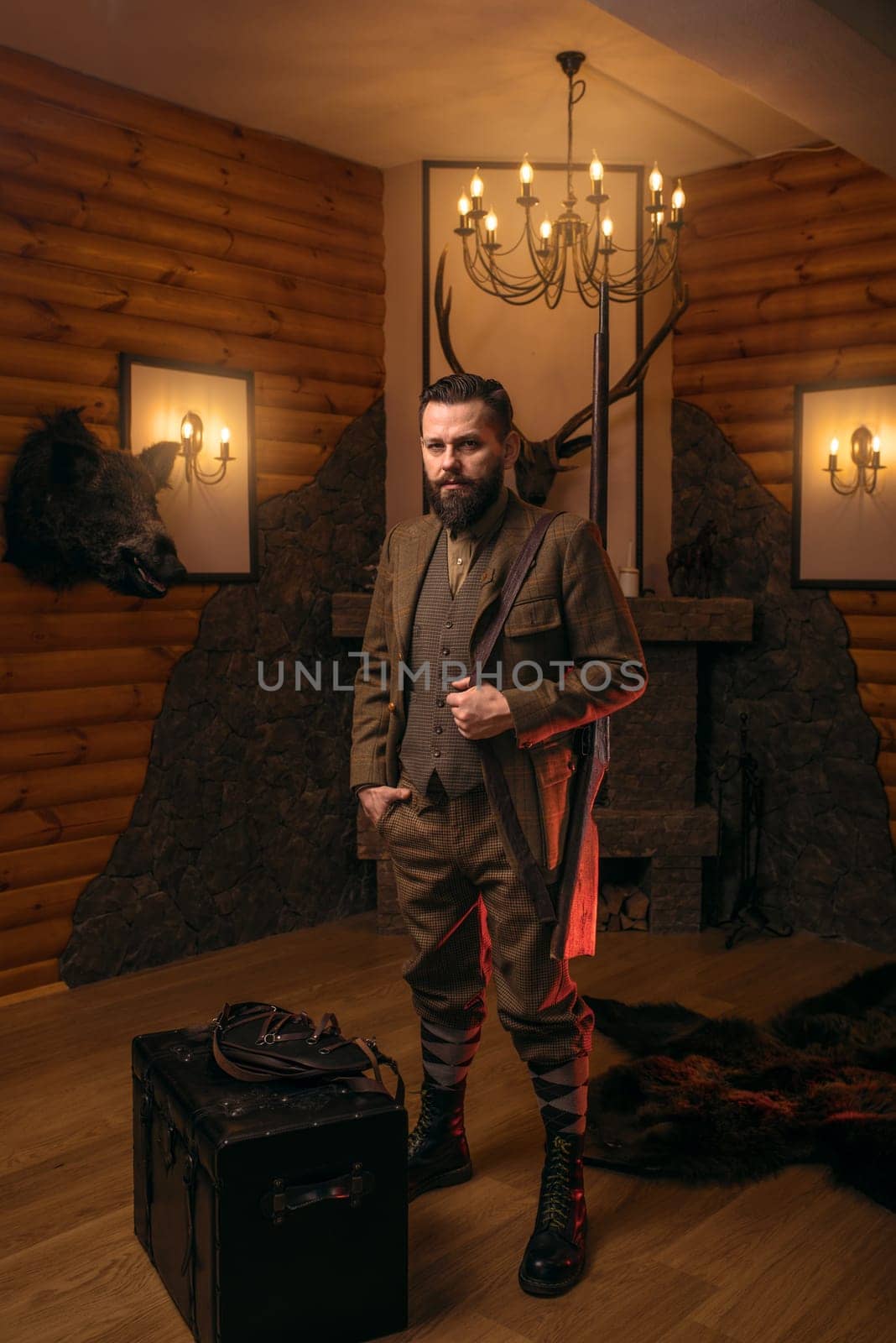 Hunter man with old gun in vintage traditional hunting clothing standing against antique chest. Fireplace, stuffed wild animals, bear skin and other trophies on background