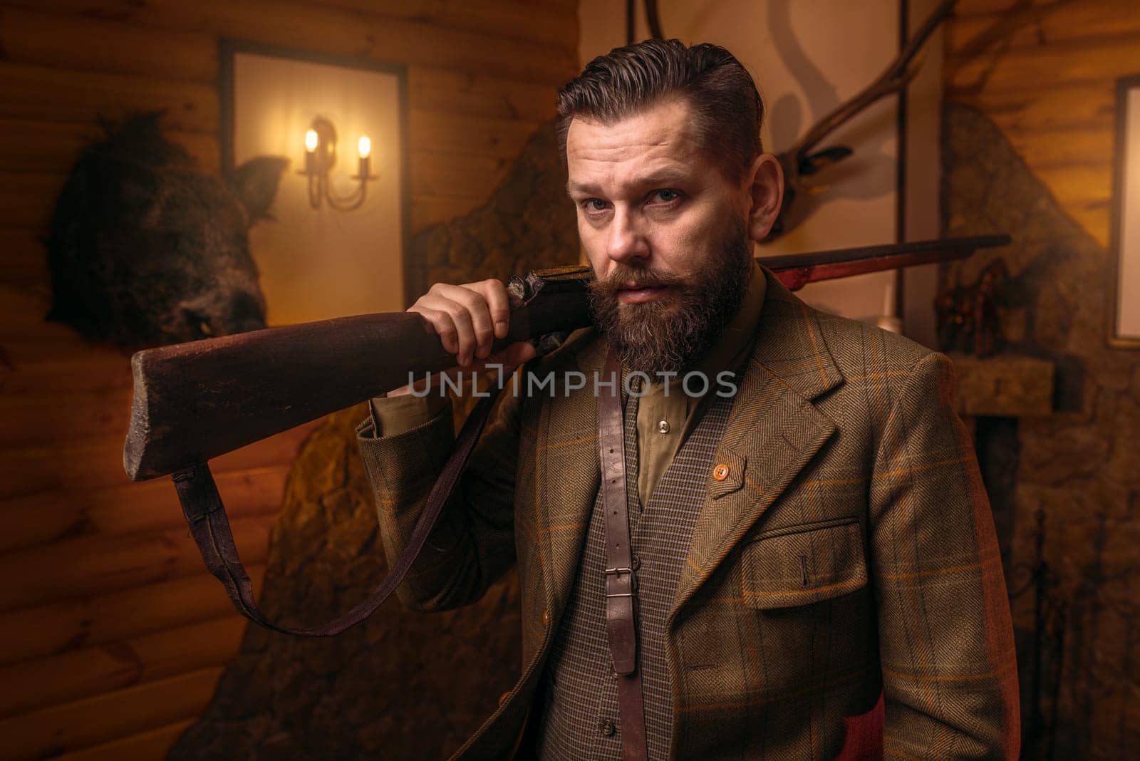 Respectable hunter man in vintage stylish hunting clothing with antique rifle against fireplace. Stuffed wild animals, bear skin and other trophies on background. Hunt lifestyle