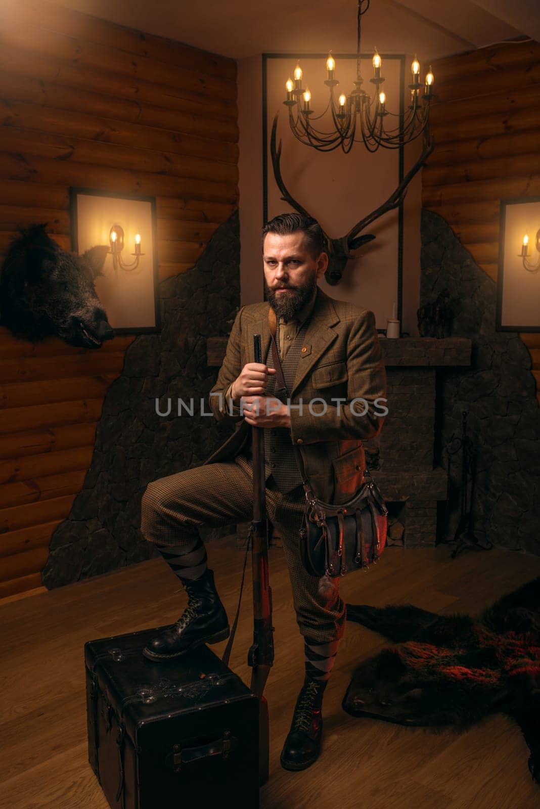Hunter man with old gun in vintage traditional hunting clothing standing against antique chest. Fireplace, stuffed wild animals, bear skin and other trophies on background