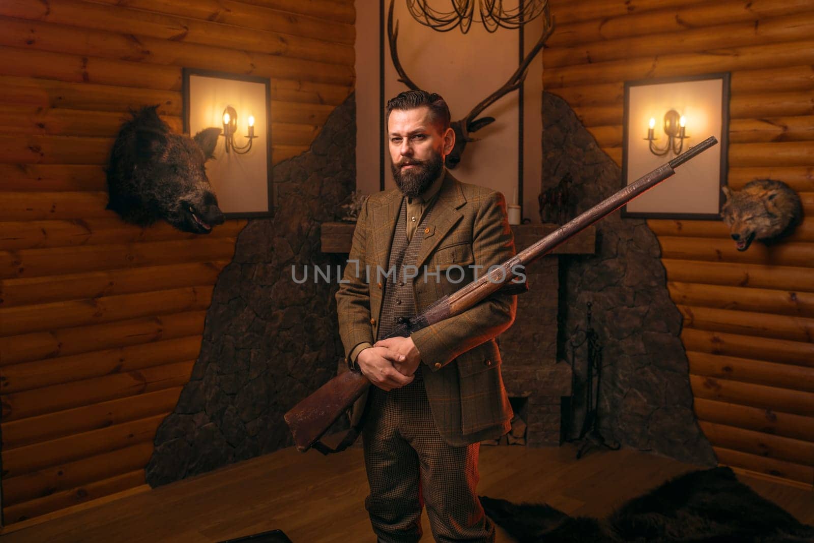 Hunter gentleman in traditional hunting clothing with old gun against fireplace. Stuffed wild animals, bear skin and other trophies on background. Old-fashioned style