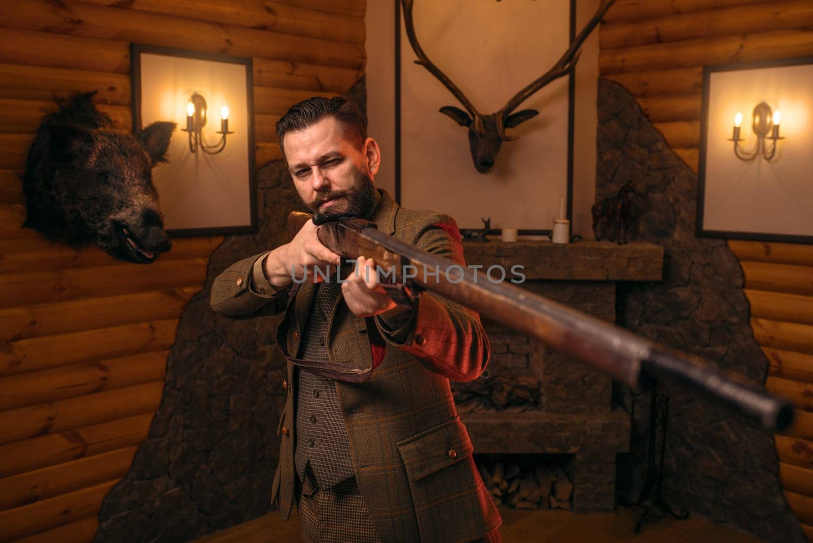 Hunter man aims of the antique hunting rifle. Fireplace, stuffed wild animals, bear skin and other trophies on background