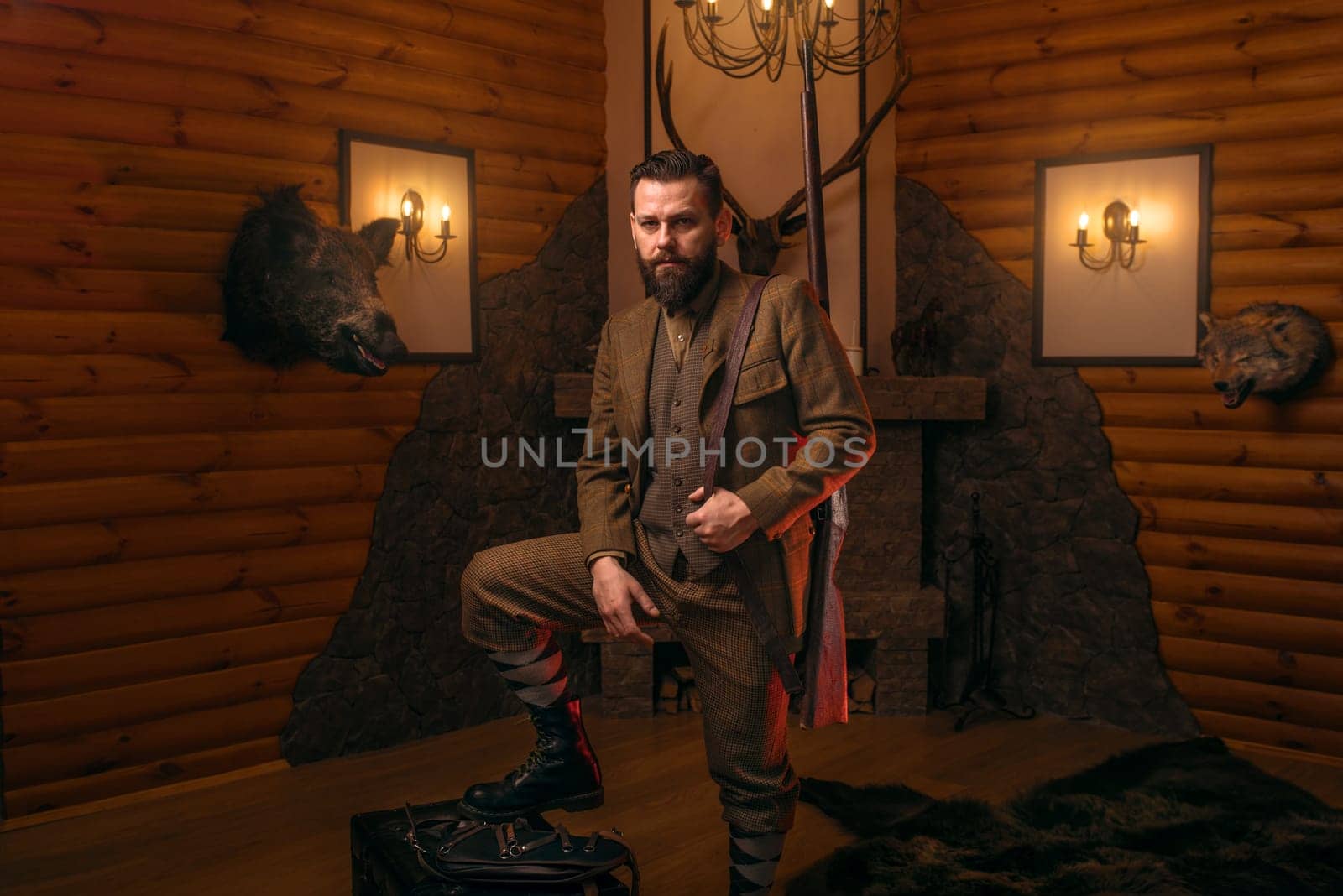Respectable hunter man with old gun in retrro style traditional hunting clothing standing against antique chest. Retro rifle, vintage clothes. Room with fireplace, stuffed wild animals, bear skin and other trophies on background