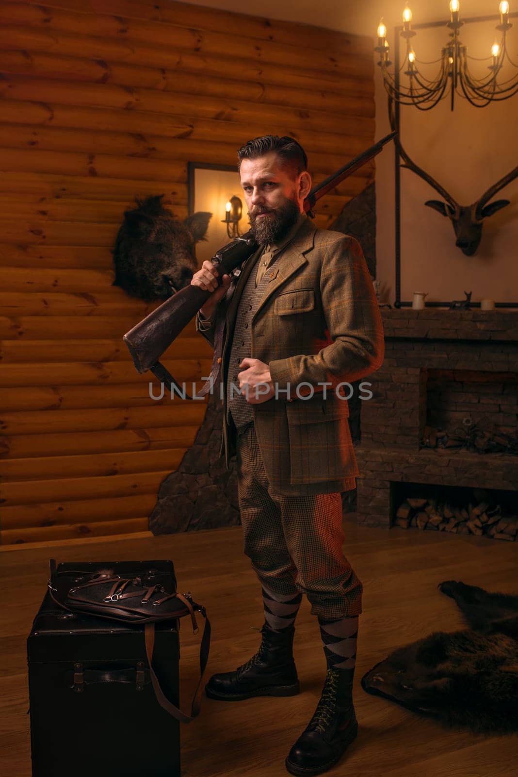 Respectable hunter man in vintage stylish hunting clothing with antique rifle against fireplace. Stuffed wild animals, bear skin and other trophies on background. Hunt lifestyle