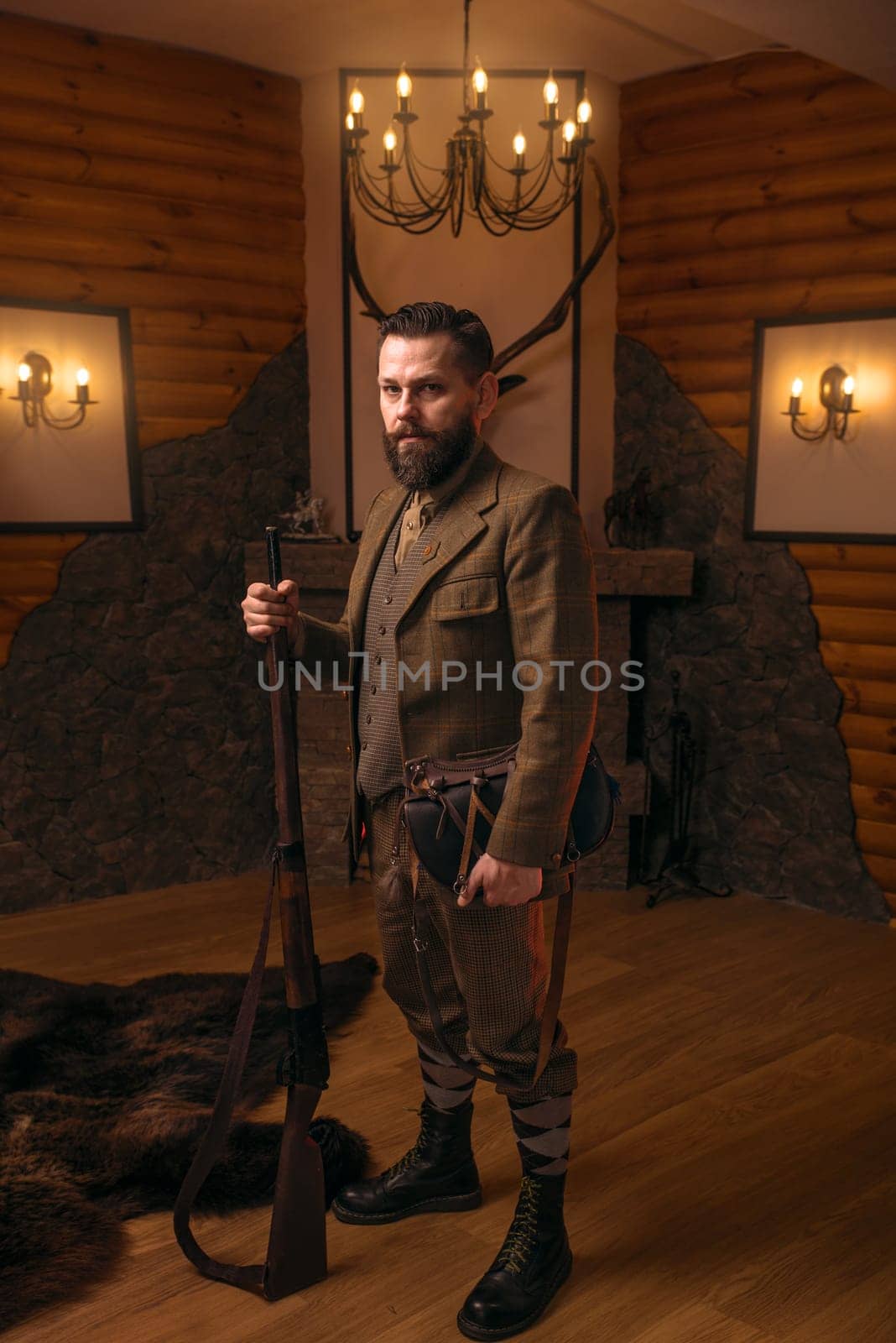 Solid hunter man in old-fashioned hunting clothing with antique gun against fireplace. Stuffed wild animals, bear skin and other trophies on background. Hunt lifestyle