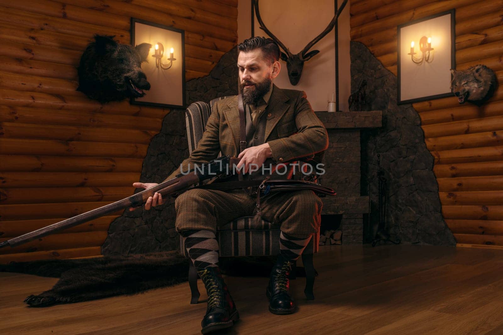 Hunter man in vintage hunting clothing sitting in a chair with antique rifle. Fireplace, stuffed wild animals, bear skin and other trophies on background. Hunt lifestyle