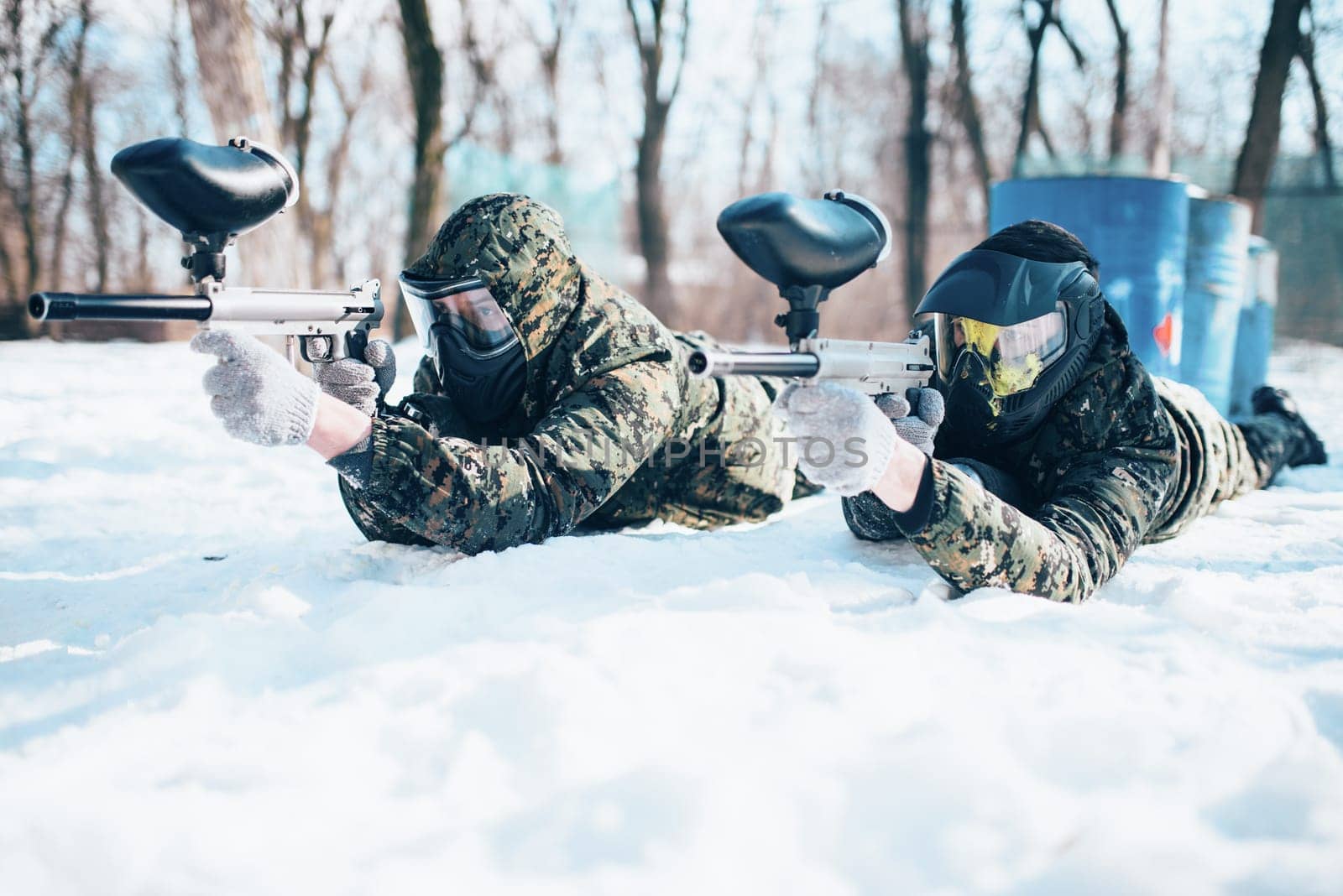 Two paintball players lies on the snow and shooting at the enemy in winter forest battle. Extreme sport game