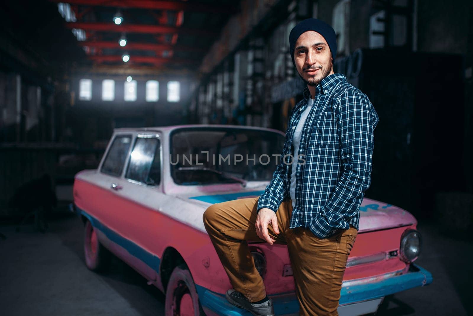 Stalker, alone traveler pose against USSR retro car, old factory interior on background. Danger zone, mysterious adventure in deserted place