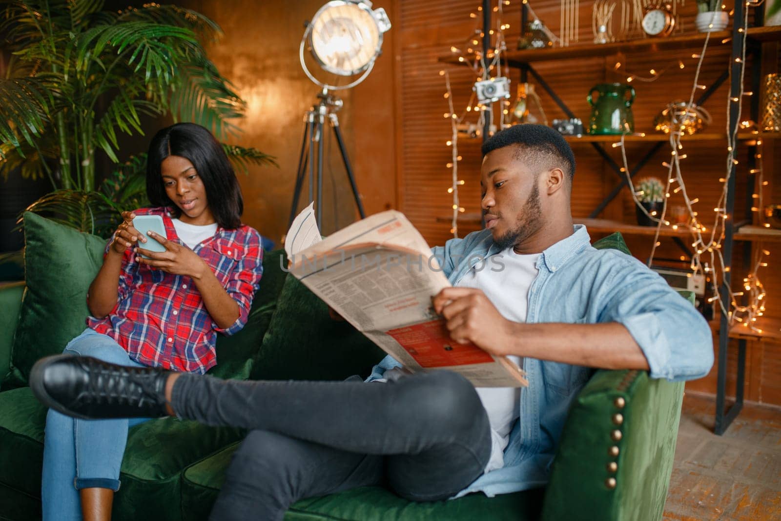Black man reading newspaper, his woman using mobile phone on couch, relaxation at home. Happy african love couple leisures in their house