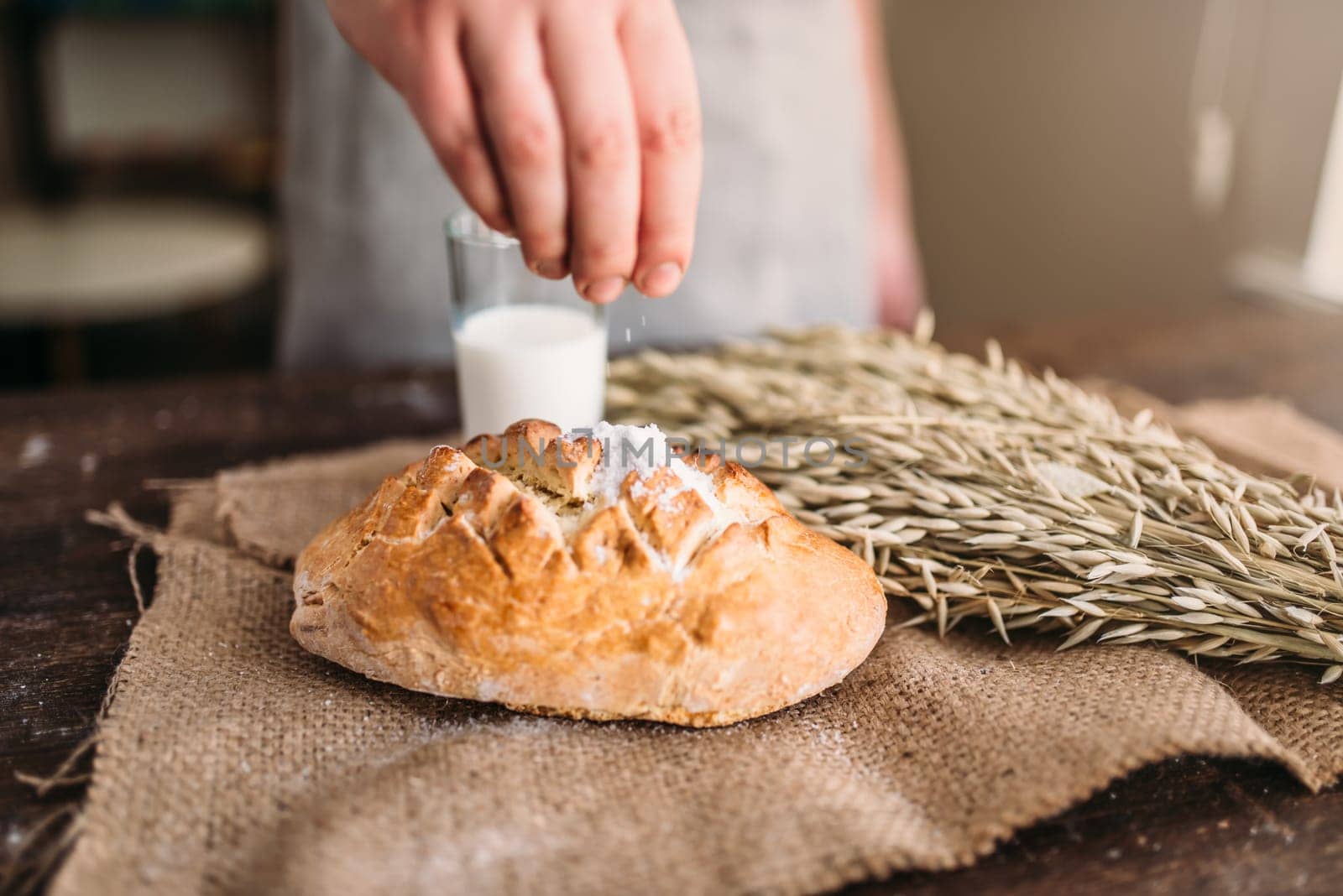 Homemade bakery concept, natural organic food. Baker hands against fresh baked bread bun with crispy crust, wheat bunch and glass of milk on burlap cloth.