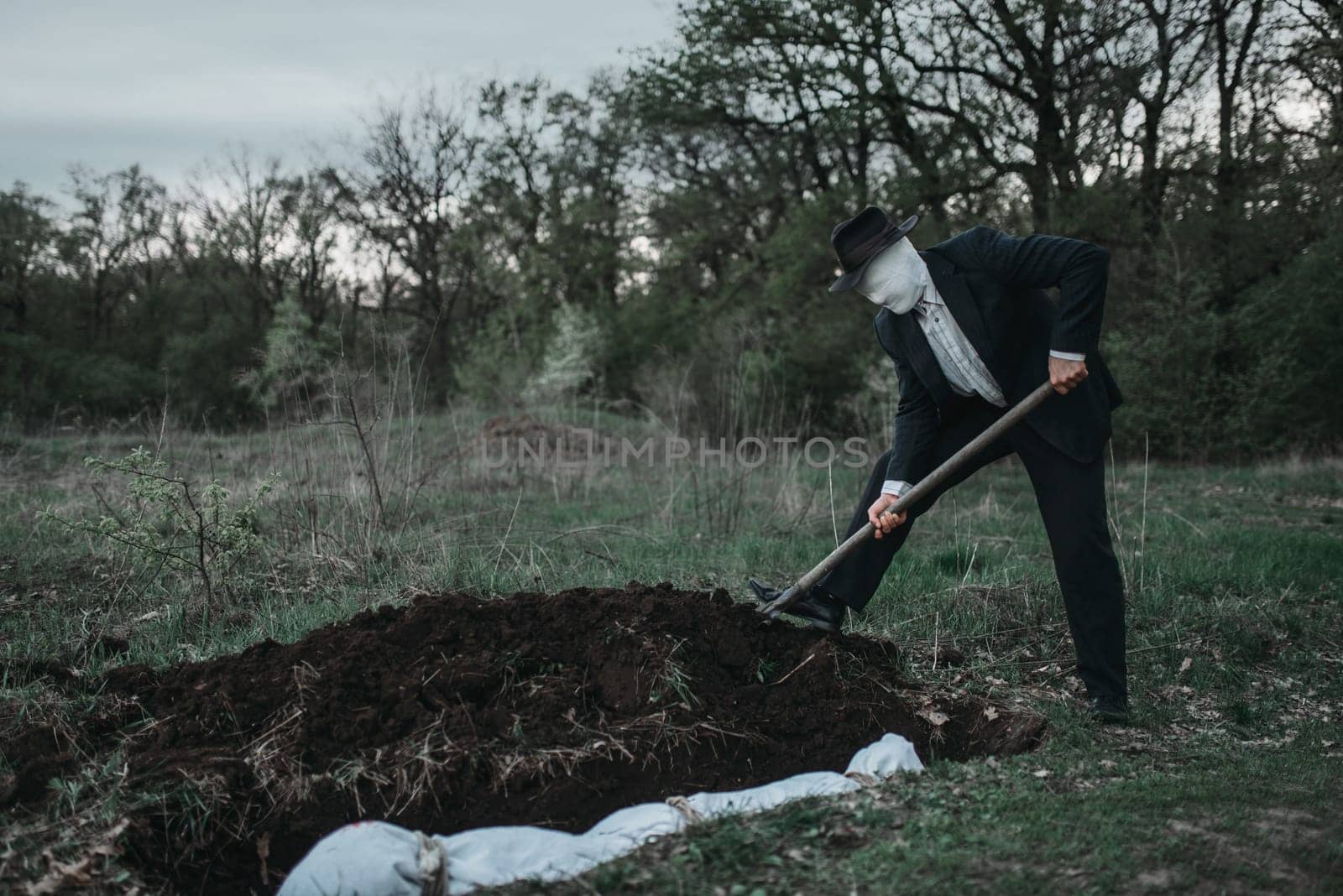 Bloody murderer is digging a grave for the victim in forest, the body wrapped in a canvas, serial maniac concept