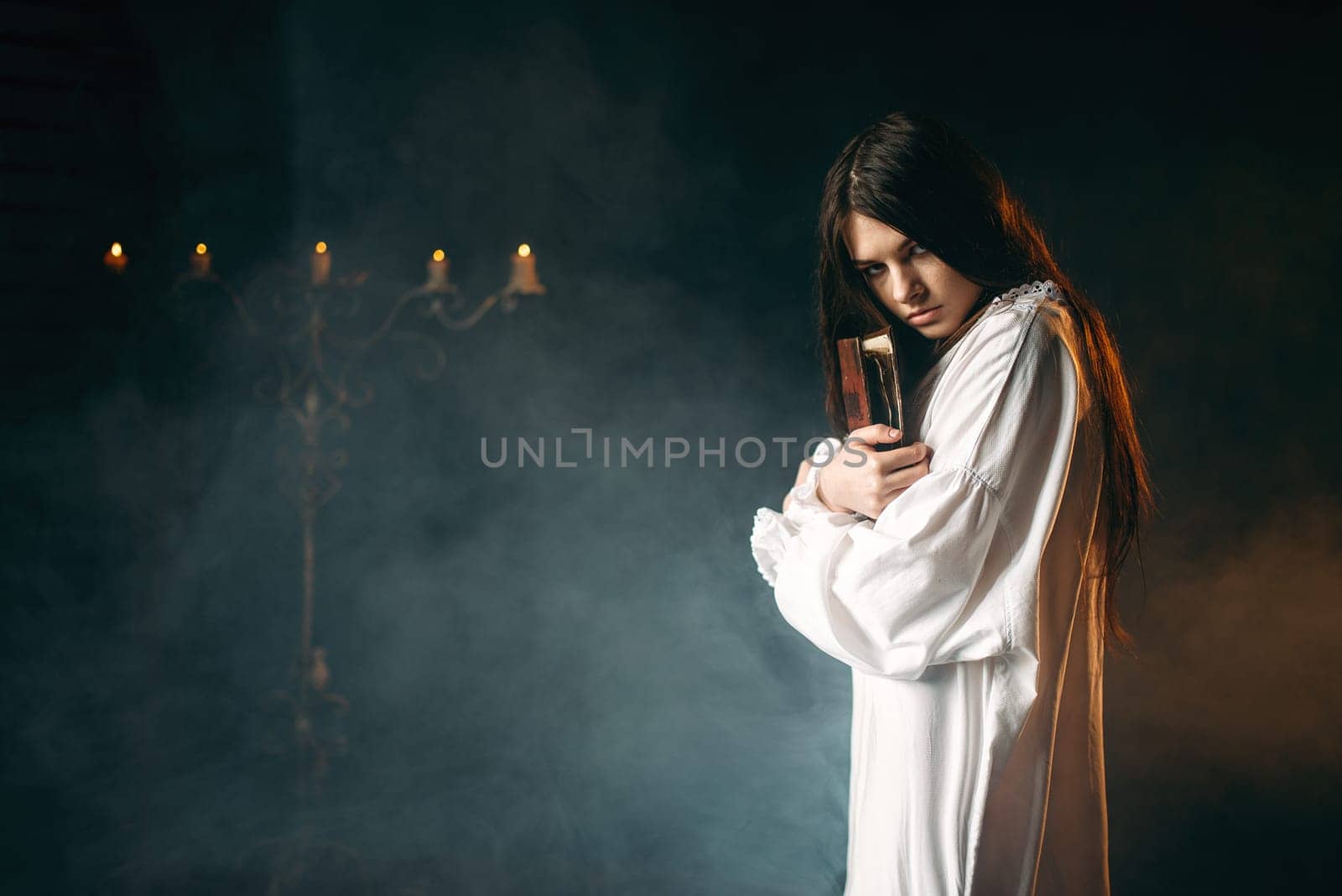 Woman in white shirt holds spellbook in hands, candles and smoke on background. Dark magic ritual, occultism and exorcism, witchcraft