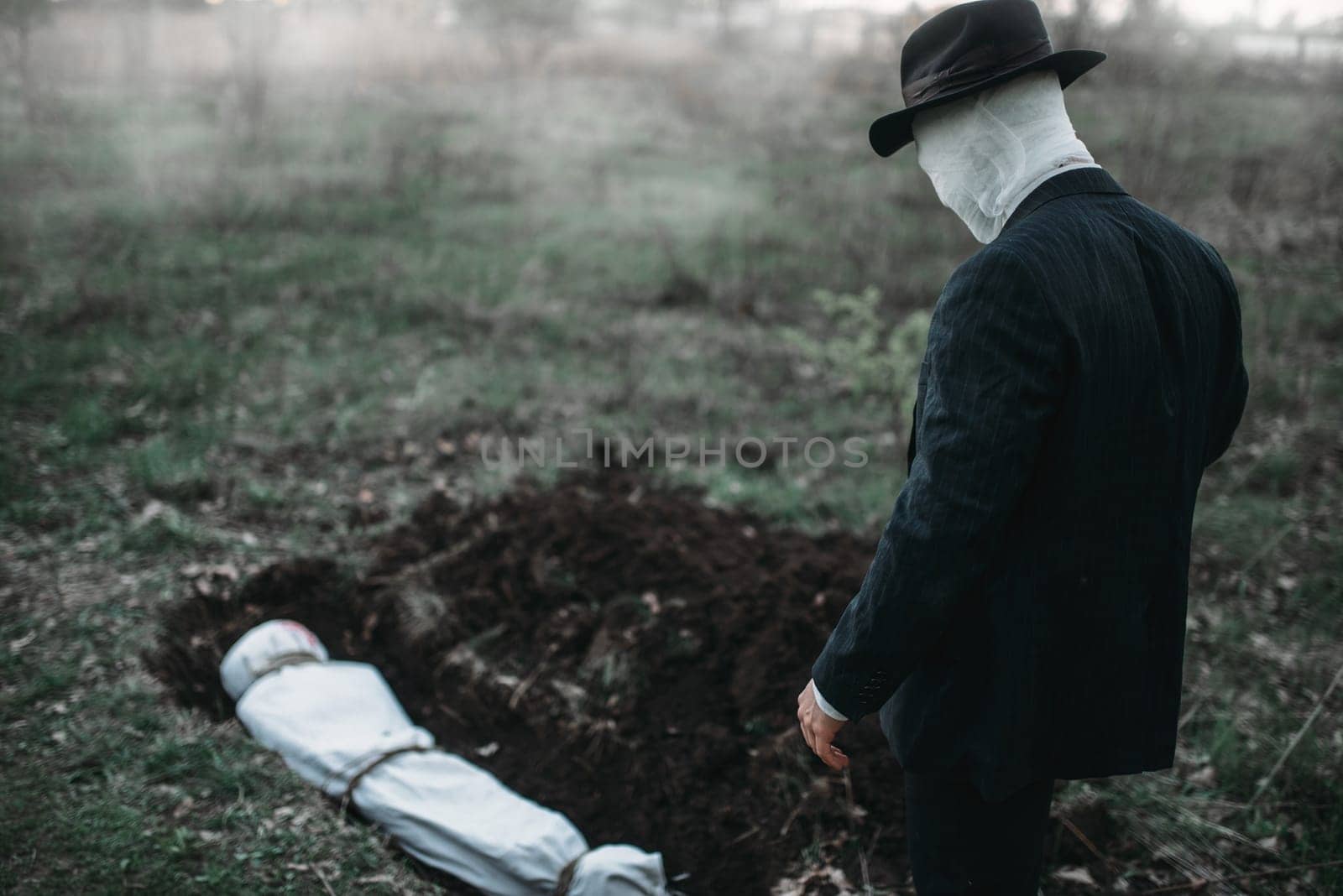 Bloody murderer stands against a grave with victim in forest, the body wrapped in a canvas, serial maniac concept