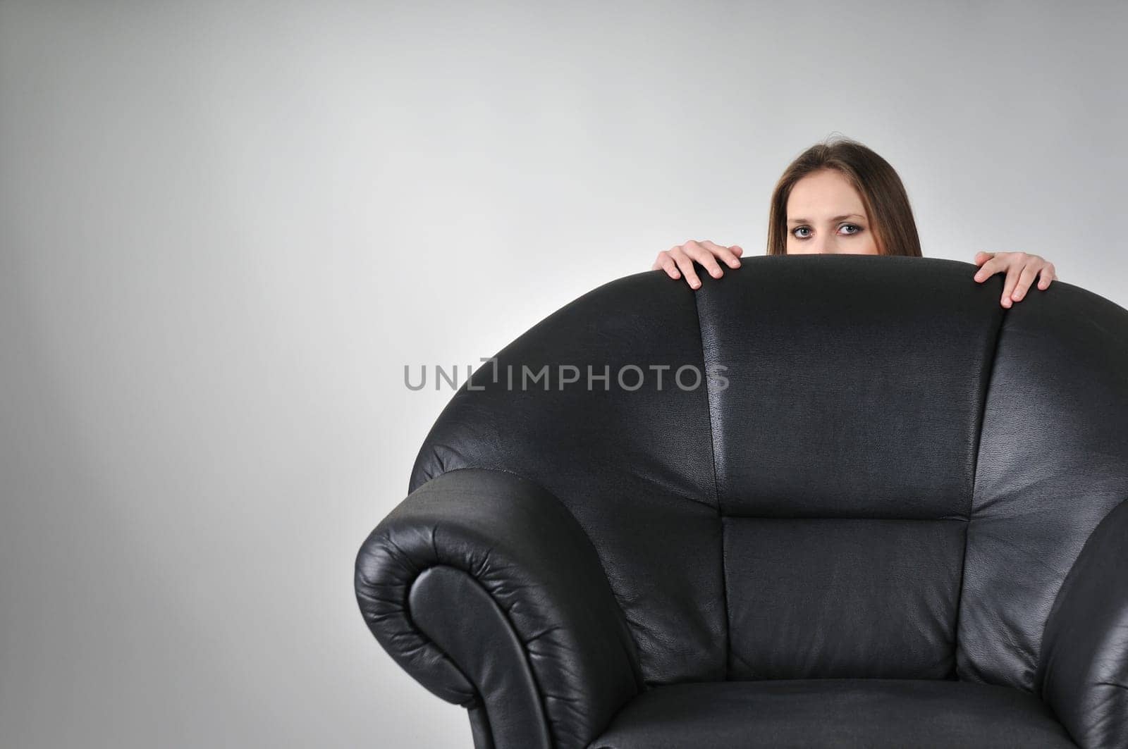 Studio portrait of young woman hidden behind big black armchair - face with hands visible