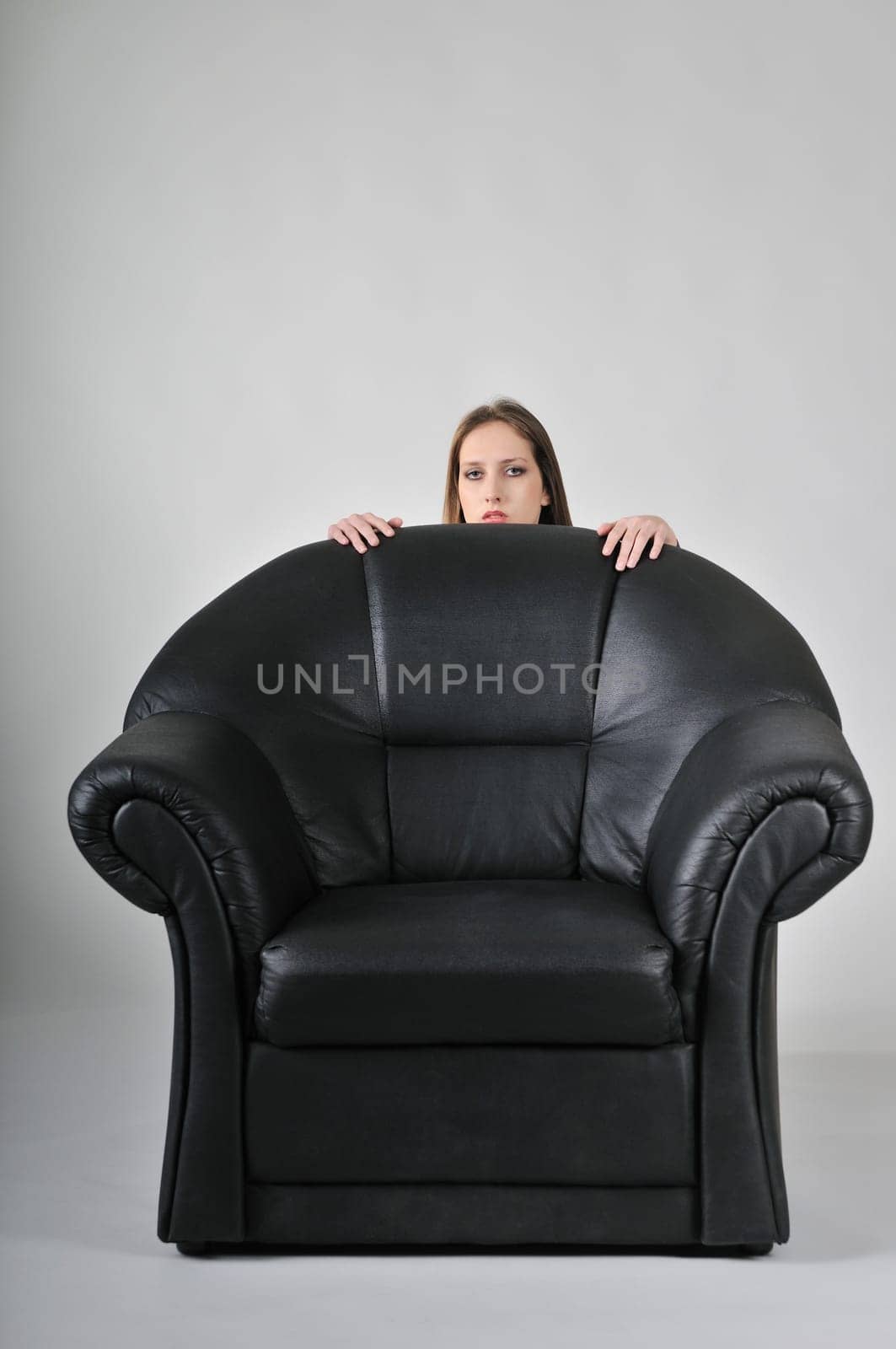 Studio portrait of young woman hidden behind big black armchair - face with hands visible