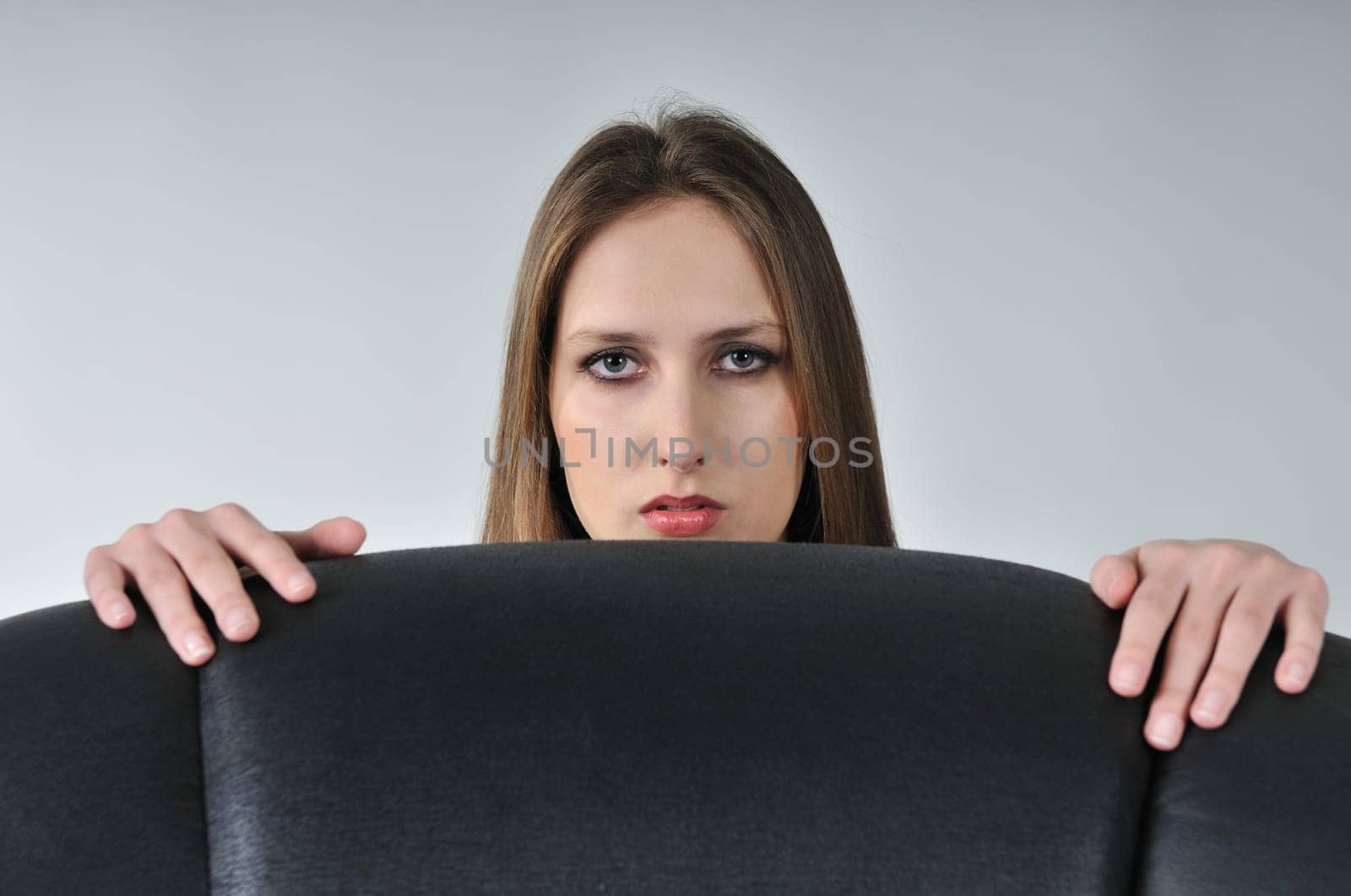 Studio portrait of young woman hidden behind big black armchair - face with hands visible