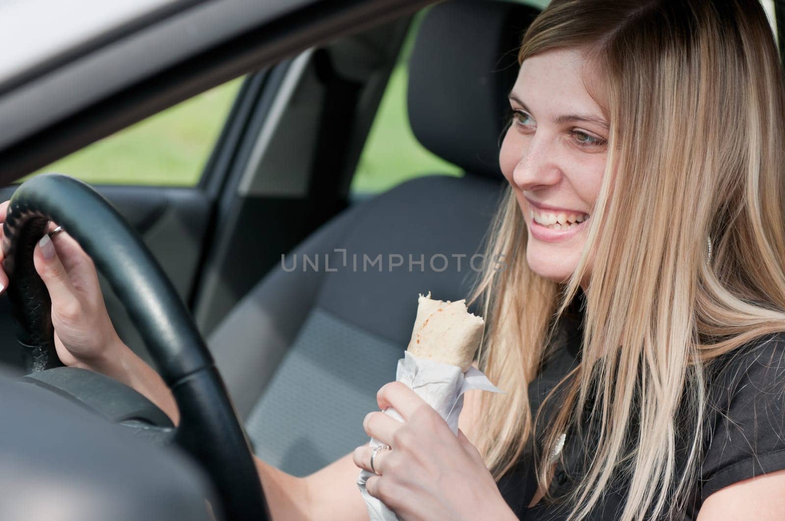 Young beautiful smiling woman driving car and eating fast food - portrait through side window