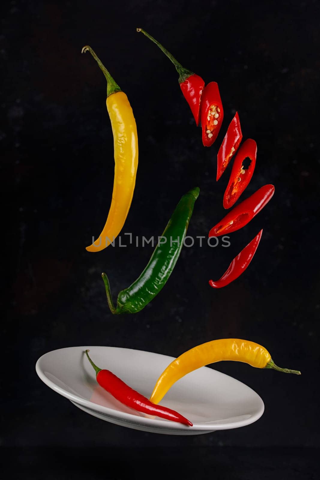 Flying red, yellow and green chili peppers on a dark background with a ceramic plate