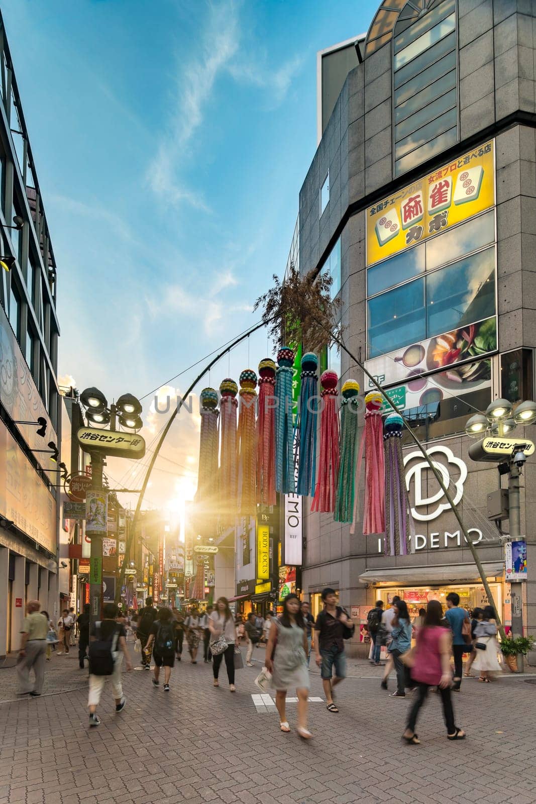 Sunset sky on Shibuya Center Gai Street which literally means Shibuya Central District. The busiest street once intersected with Shibuya Station, it stretches for 350 meters and incorporates the adjacent streets of Bunkamura, Inokashira and Utagawa into its neighborhood. In 2011 the street was renamed Basketball Street in reference to the holy place of Japanese basketball which is the Yoyogi National Gymnasium to which this street leads. The photo is taken in summer at the Tanabata Star Festival where the portal is adorned with paper decorations in the shape of shooting stars for the occasion.