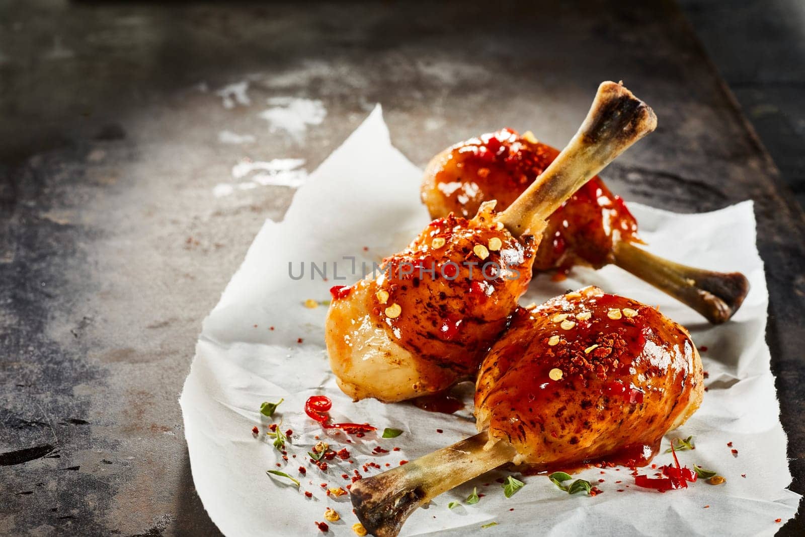 Spicy hot grilled chicken legs with chili sauce and herbs and spice seasoning served on crumpled white paper on a dark rustic background