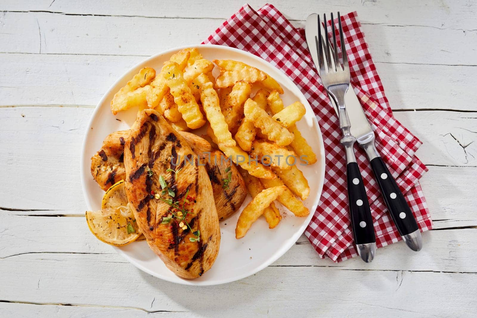 Barbecued healthy skinless chicken breast seasoned with herbs and served with crinkle cut potato chips viewed from overhead with utensils and napkin on rustic white wood