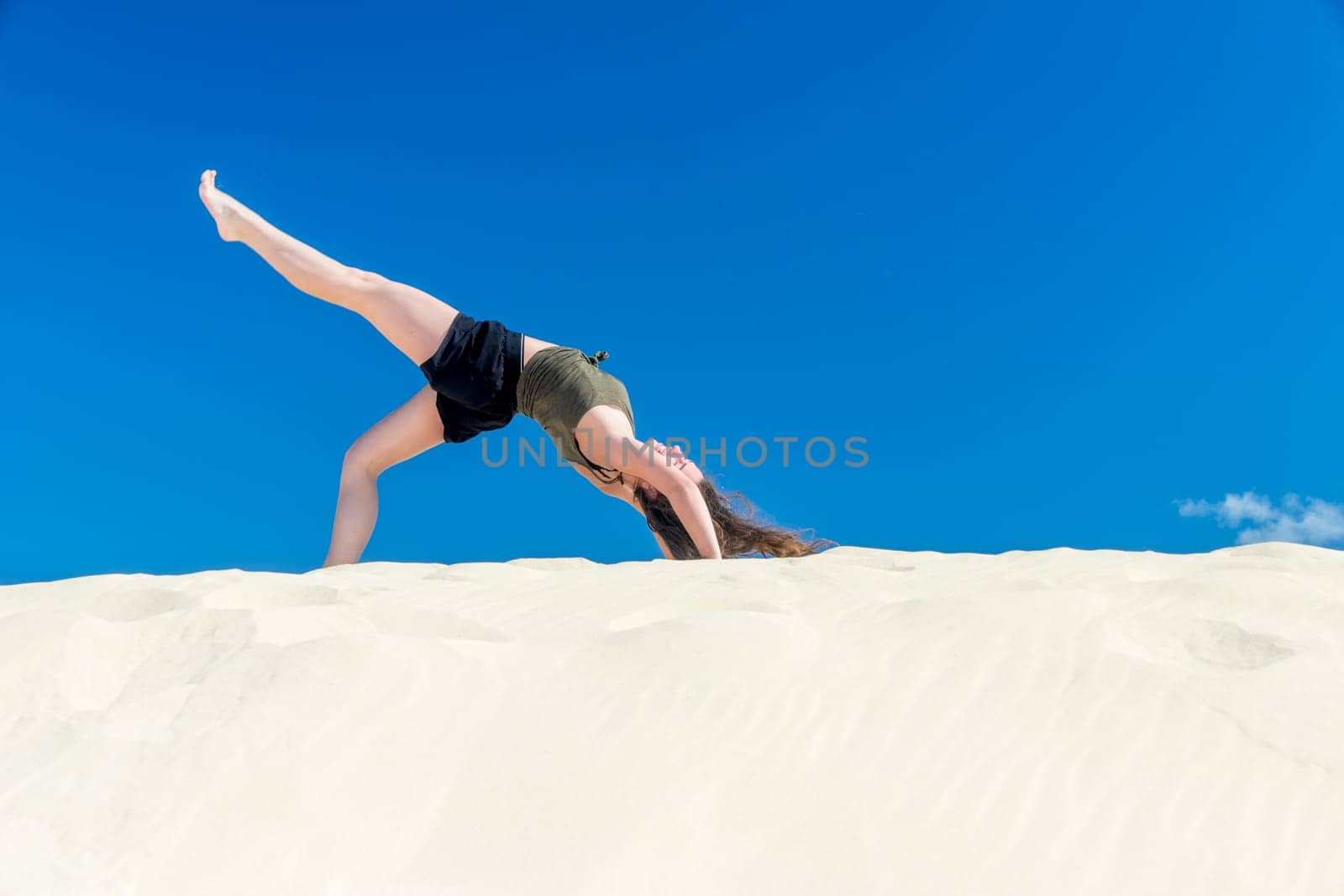 A young girl does yoga in the sand against a blue sky in summer