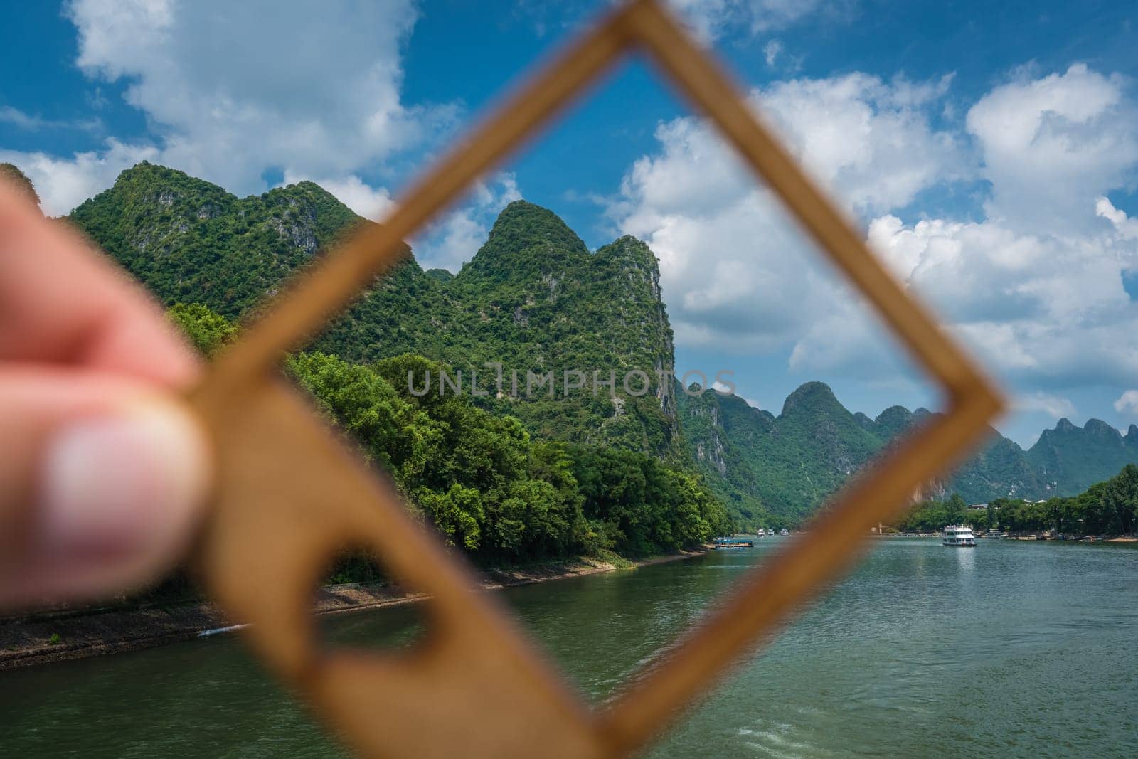 Man holding a small picture frame covering green karst peak dotting the banks of the magnificent Li river flowing from Guilin to Yangshuo, China