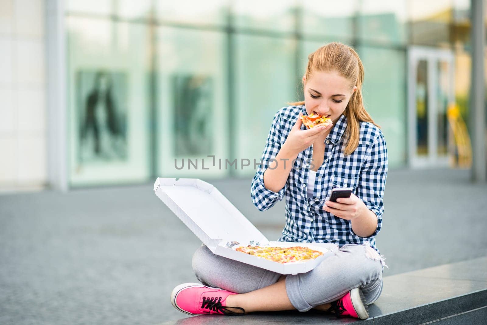Teenager eating pizza in street and browsing internet on phone