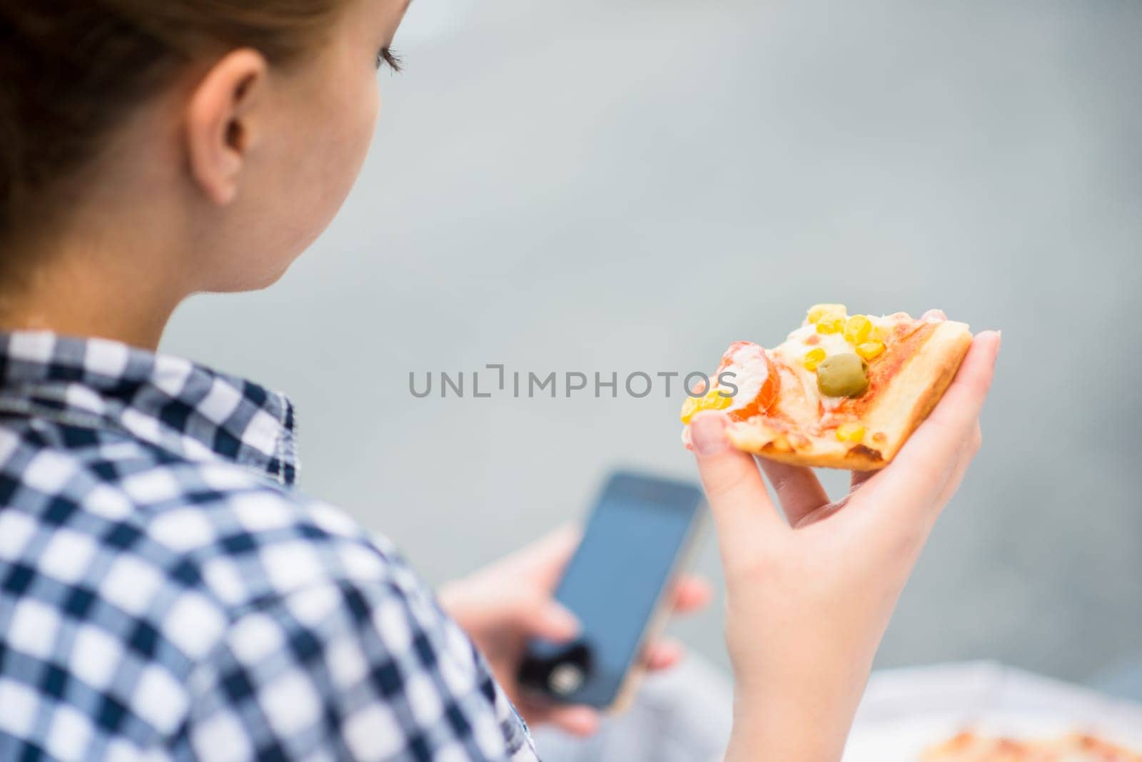 Teenager eating pizza in street and browsing internet on phone - rear view
