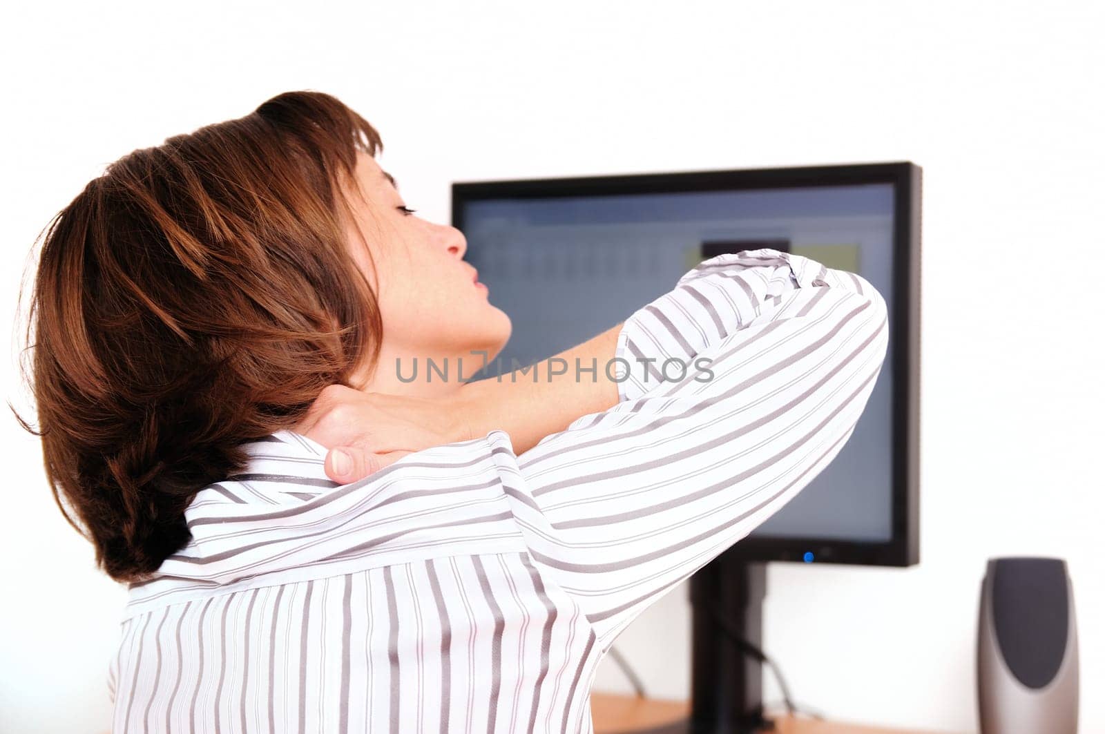 Business woman holding her neck with hand. Focus on hand on neck with blurred monitor on table in background.