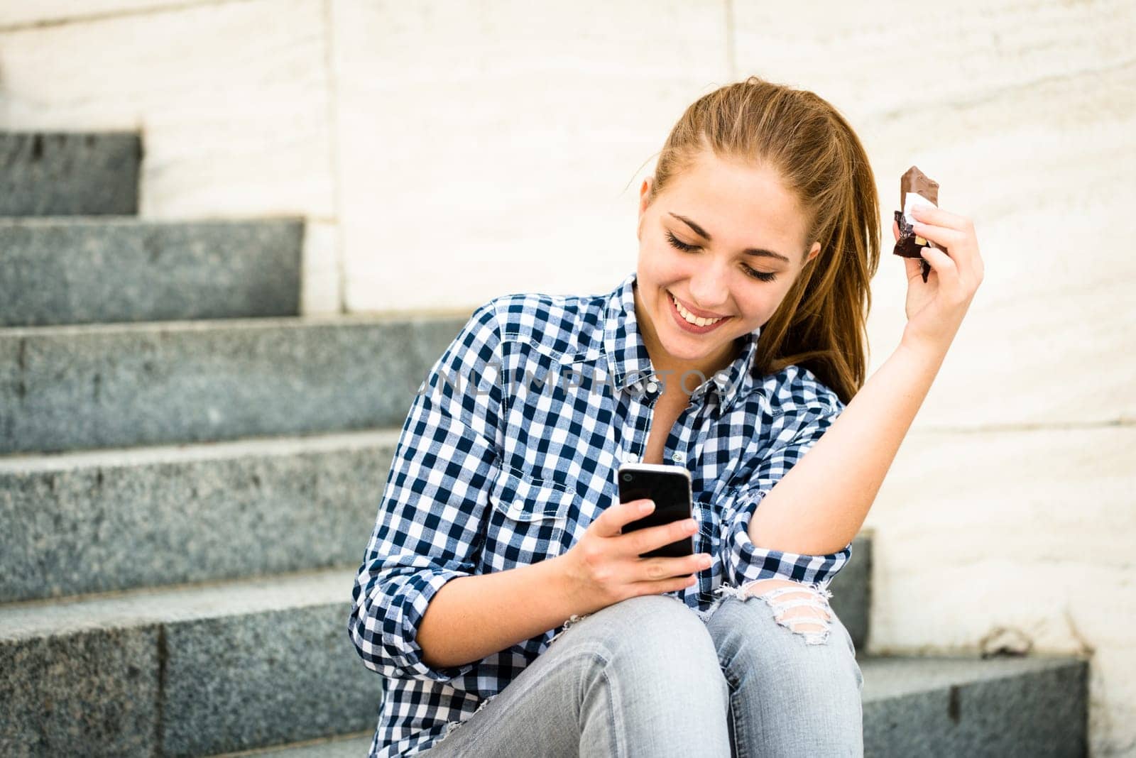 Teenager - young woman eating chocolate in street and looking in phone