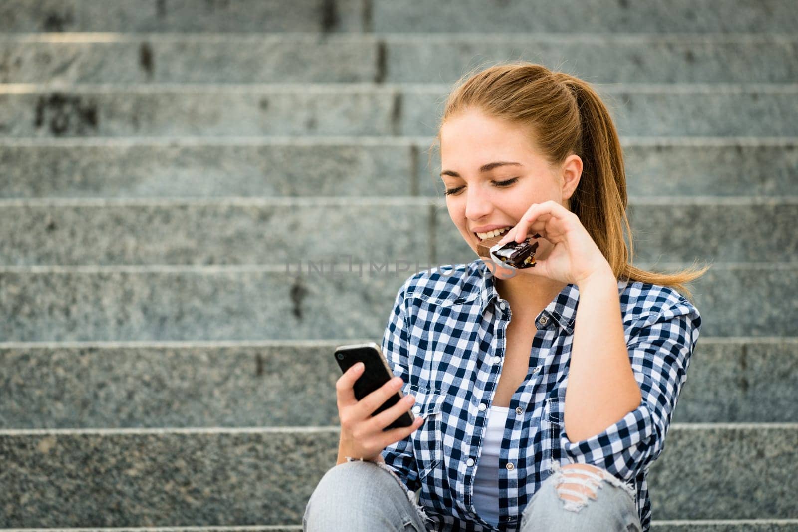 Teenager - young woman eating chocolate in street and looking in phone