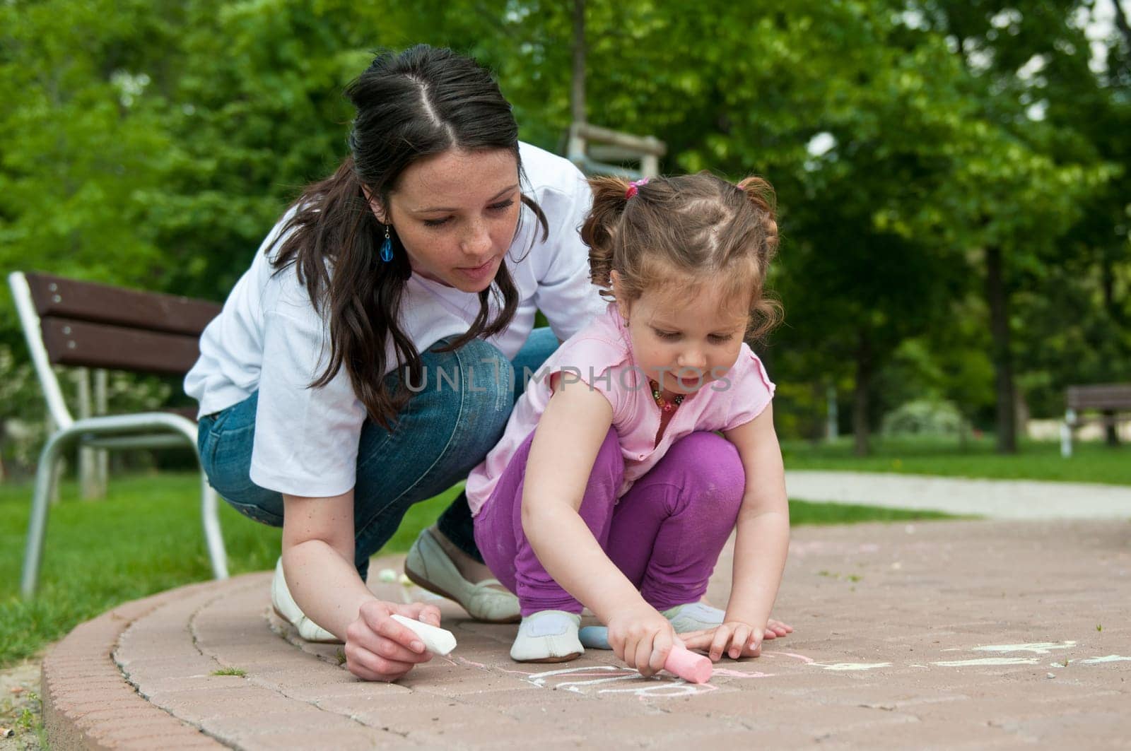 Mother is teaching her cute daughter to draw with chalk on sidewalk