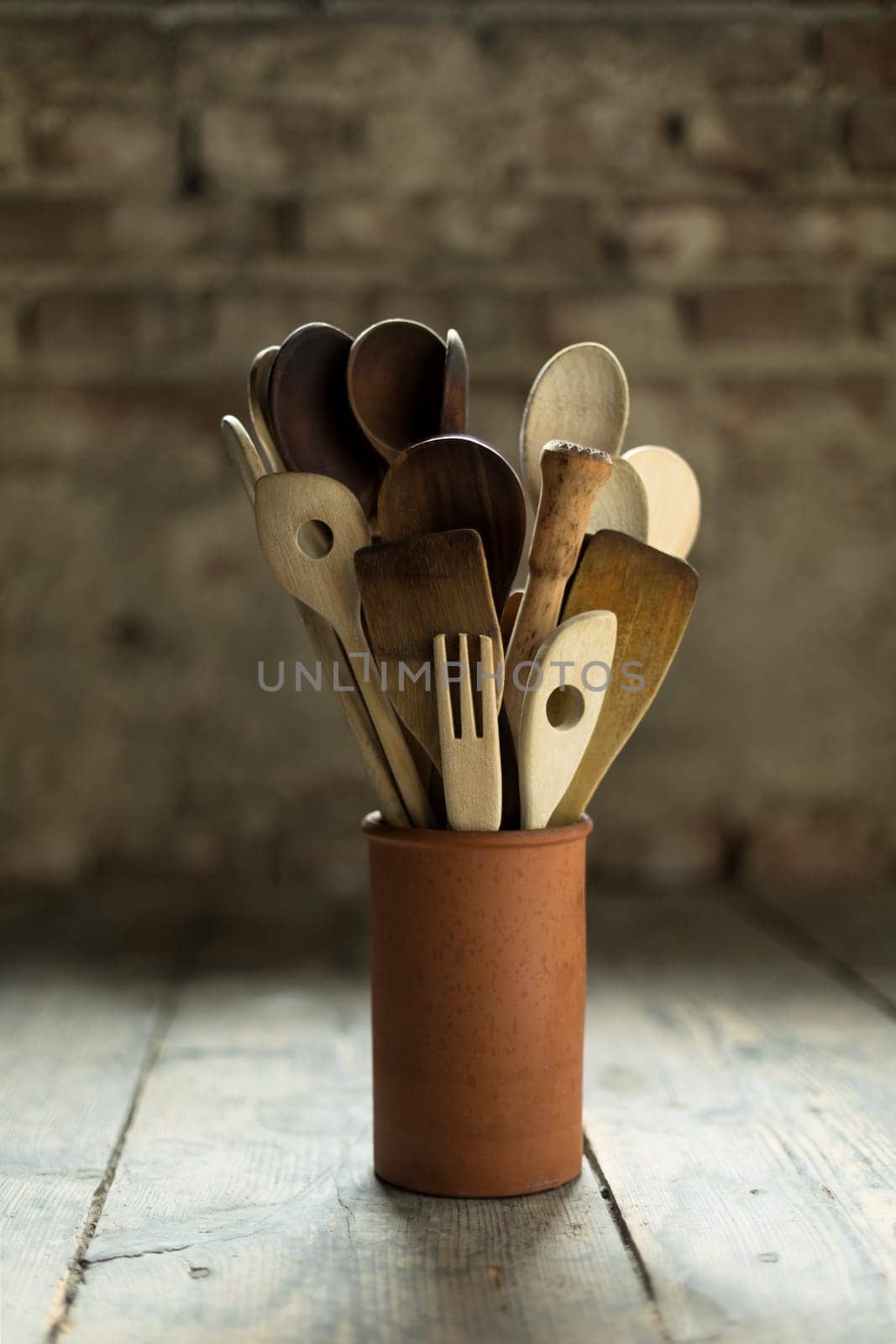 old wooden spoons in a clay vase on a deal board floor in front of a brick wall