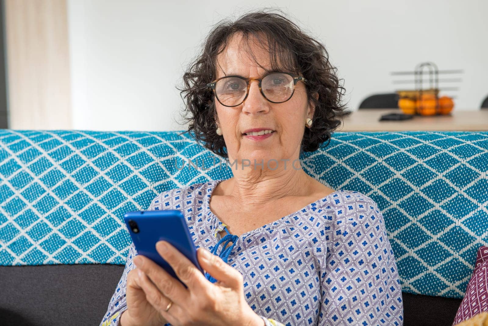 Portrait of cheerful brunette senior woman using smartphone while sitting on sofa