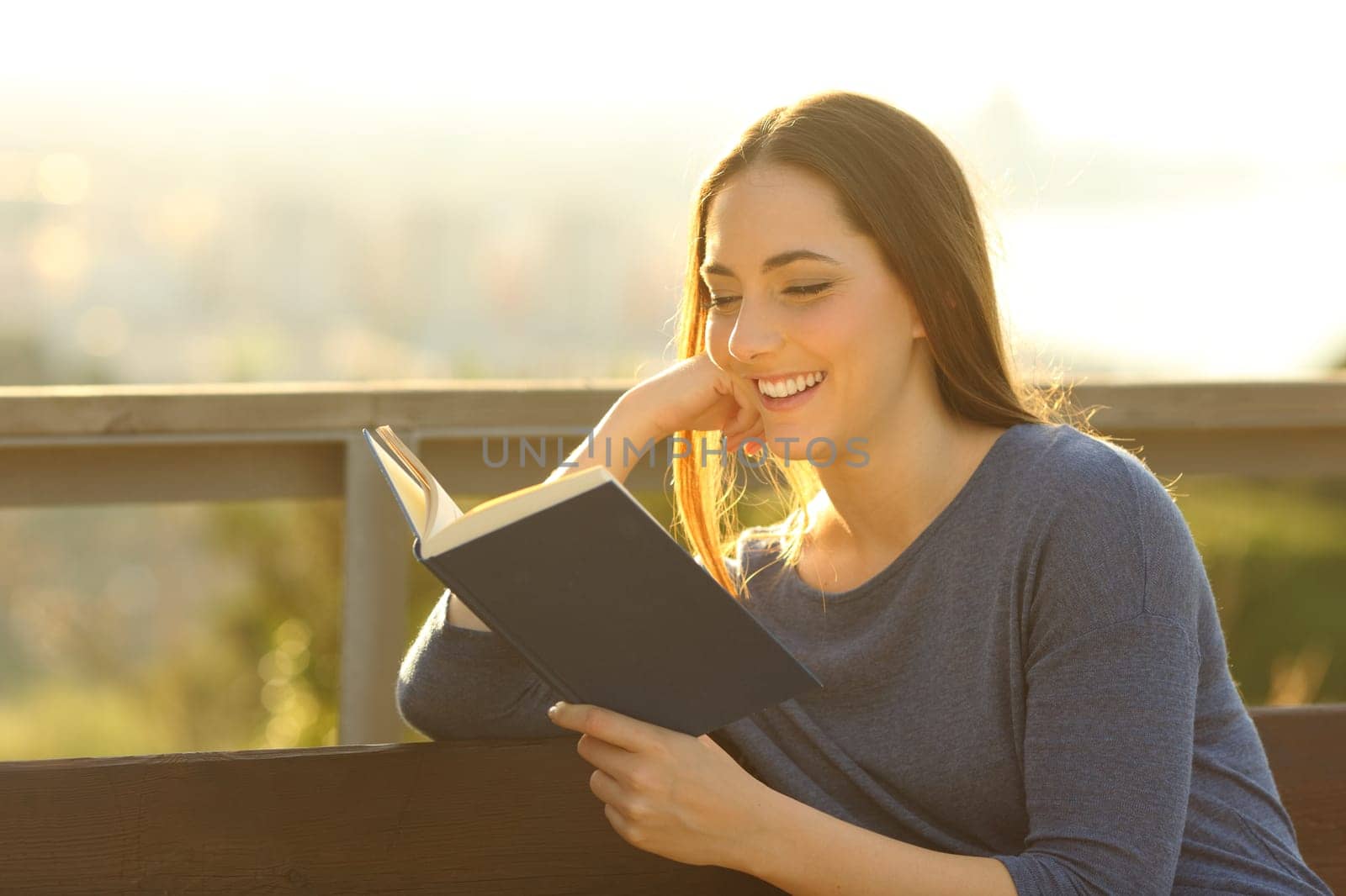 Happy woman reading an interesting book and sitting on a bench outdoors at sunset